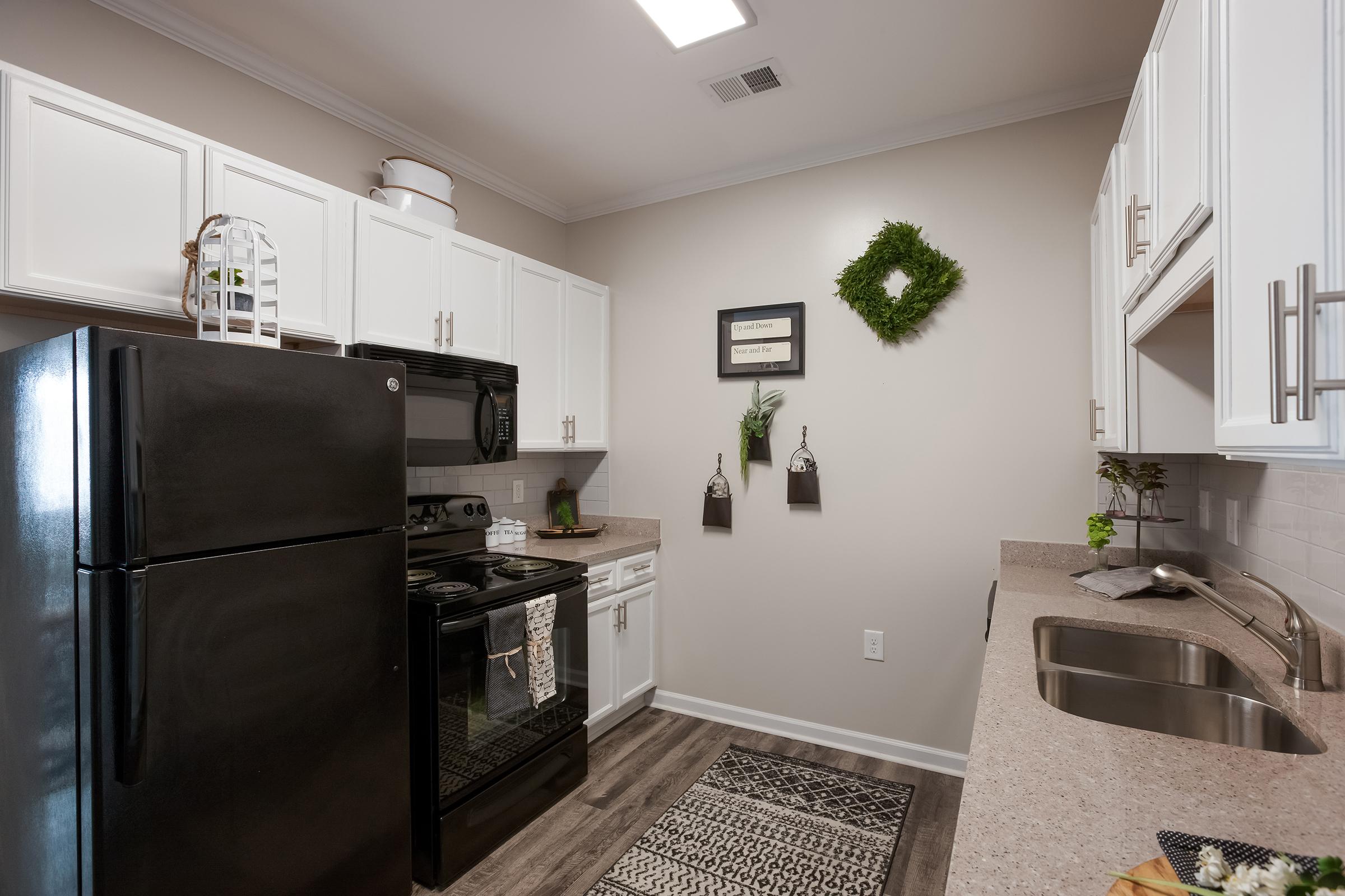 A modern kitchen featuring white cabinetry, black appliances, and a gray countertop. The room includes a refrigerator, stove, and sink, with decorative items like wall art and greenery. The flooring is a dark wood-look laminate, giving the space a cozy and inviting atmosphere.