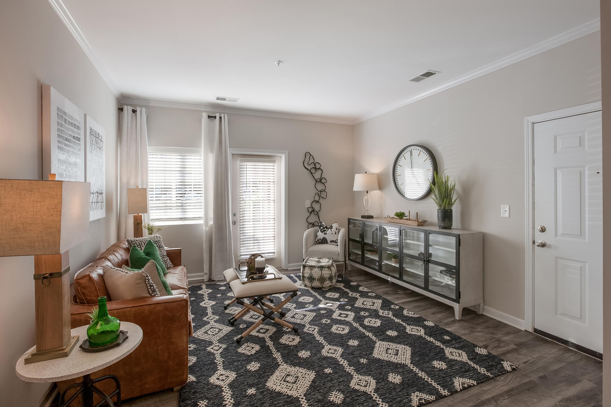 A cozy living room featuring a dark sofa with decorative pillows, two patterned armchairs, a colorful area rug, and a wooden side table with a lamp. The room has several windows providing natural light and views of greenery outside, complemented by warm wall colors and framed artwork.