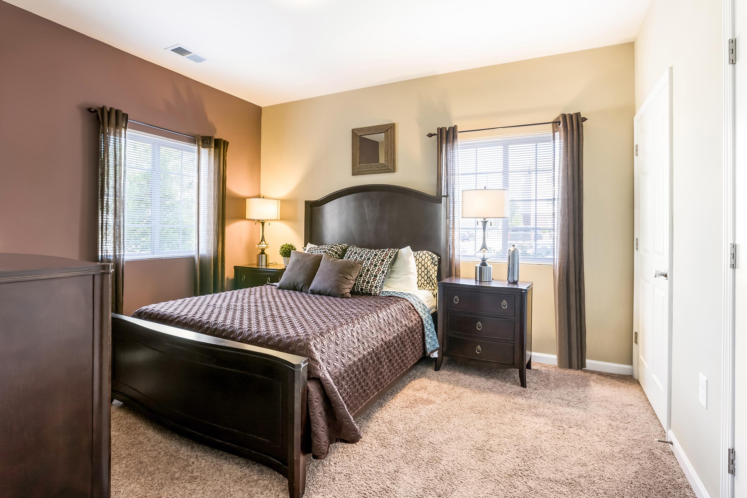 A cozy bedroom featuring a queen-sized bed with dark bedding, two bedside lamps, and a coordinating nightstand. The walls are painted in warm colors, with large windows allowing natural light to brighten the space. A dresser is visible, and plush carpet covers the floor, enhancing the room's comfort.