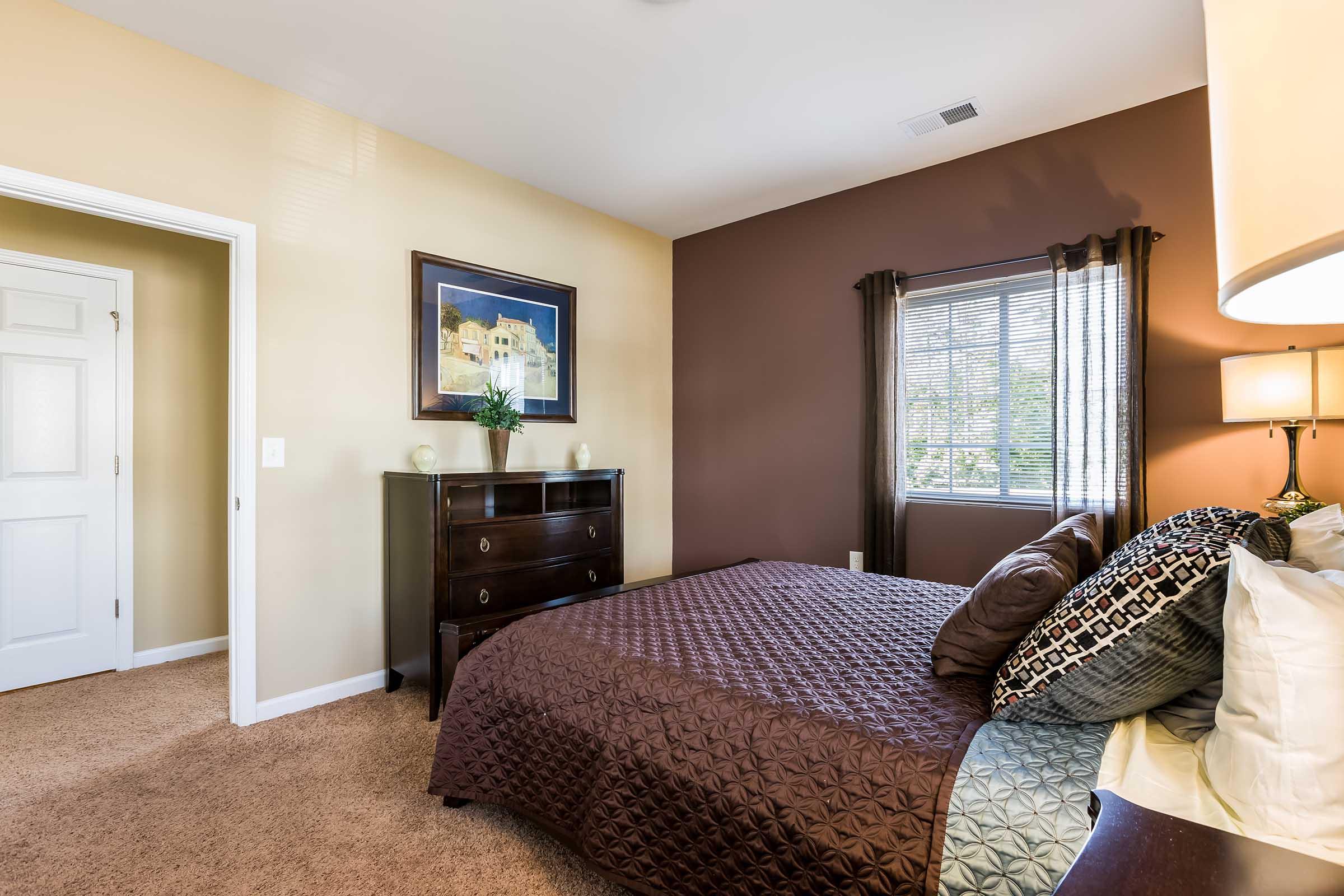 A cozy bedroom featuring a queen-sized bed with a decorative brown and cream quilt. There are accent pillows on the bed, a wooden dresser against the wall, and a framed picture above it. A window with curtains allows natural light in, and the walls are painted in warm tones, creating a welcoming atmosphere.
