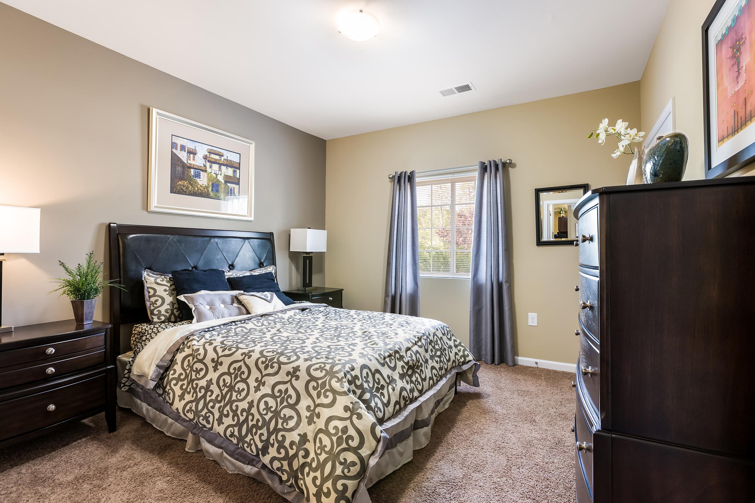 A well-decorated bedroom featuring a queen-sized bed with an ornate black headboard and patterned bedding. There are two bedside lamps on matching nightstands, a wooden dresser, and a window with gray curtains letting in natural light. Wall art and a potted plant add to the cozy ambiance.