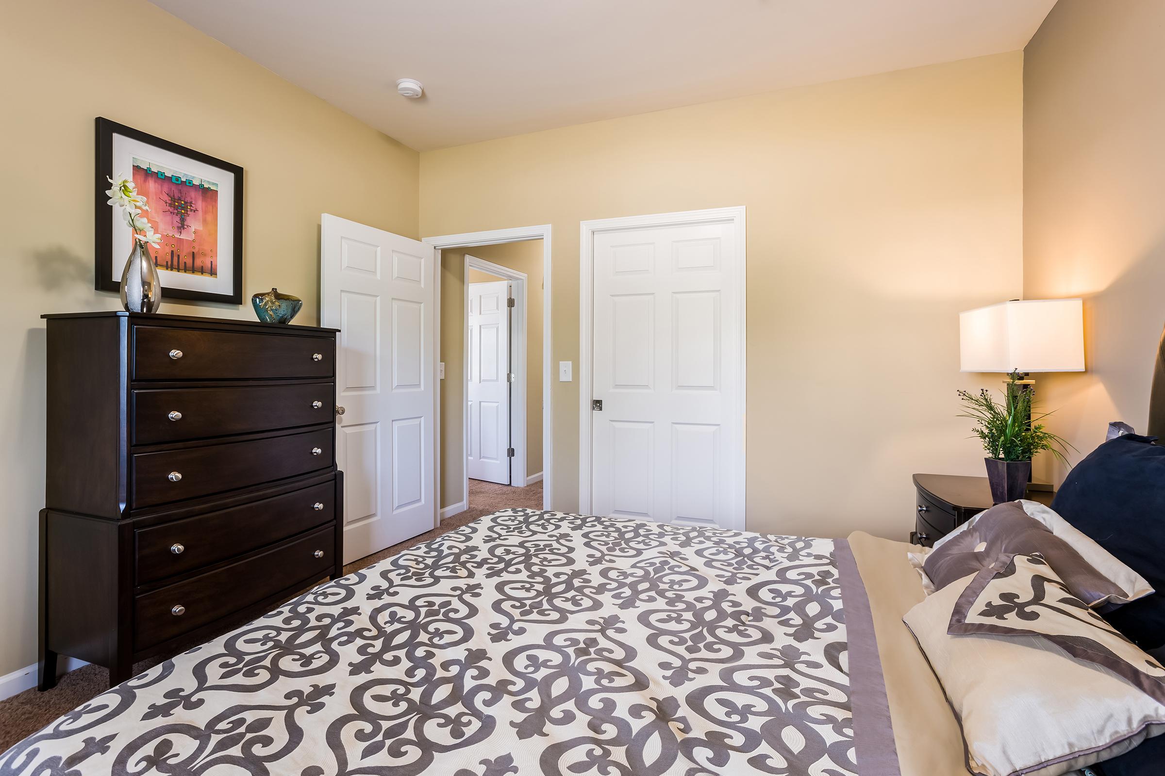 Cozy bedroom featuring a neatly made bed with decorative pillows, a dark wood dresser, and a bedside table with a lamp. The walls are painted in a soft beige, and there are two white doors leading to other rooms. A framed picture and a small plant add a touch of decor to the space.