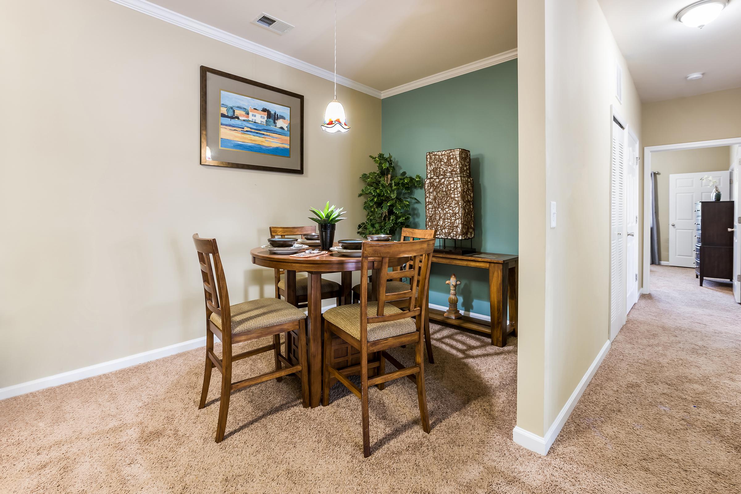 A well-decorated dining area featuring a round wooden table with four chairs, a plant centerpiece, and a framed painting on the wall. The room has light beige walls, a green accent wall, and a carpeted floor. A small console table stands against the wall, with a door visible in the background.