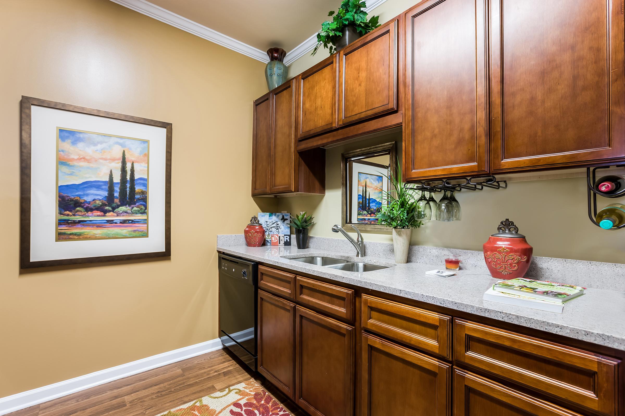 A warm-toned kitchen featuring rich wooden cabinets, a double sink, and a countertop with decorative elements. A framed landscape painting hangs on the wall, reflecting greenery and mountains. Shelves display potted plants and bottles, while a colorful rug adds a cozy touch to the wooden floor.