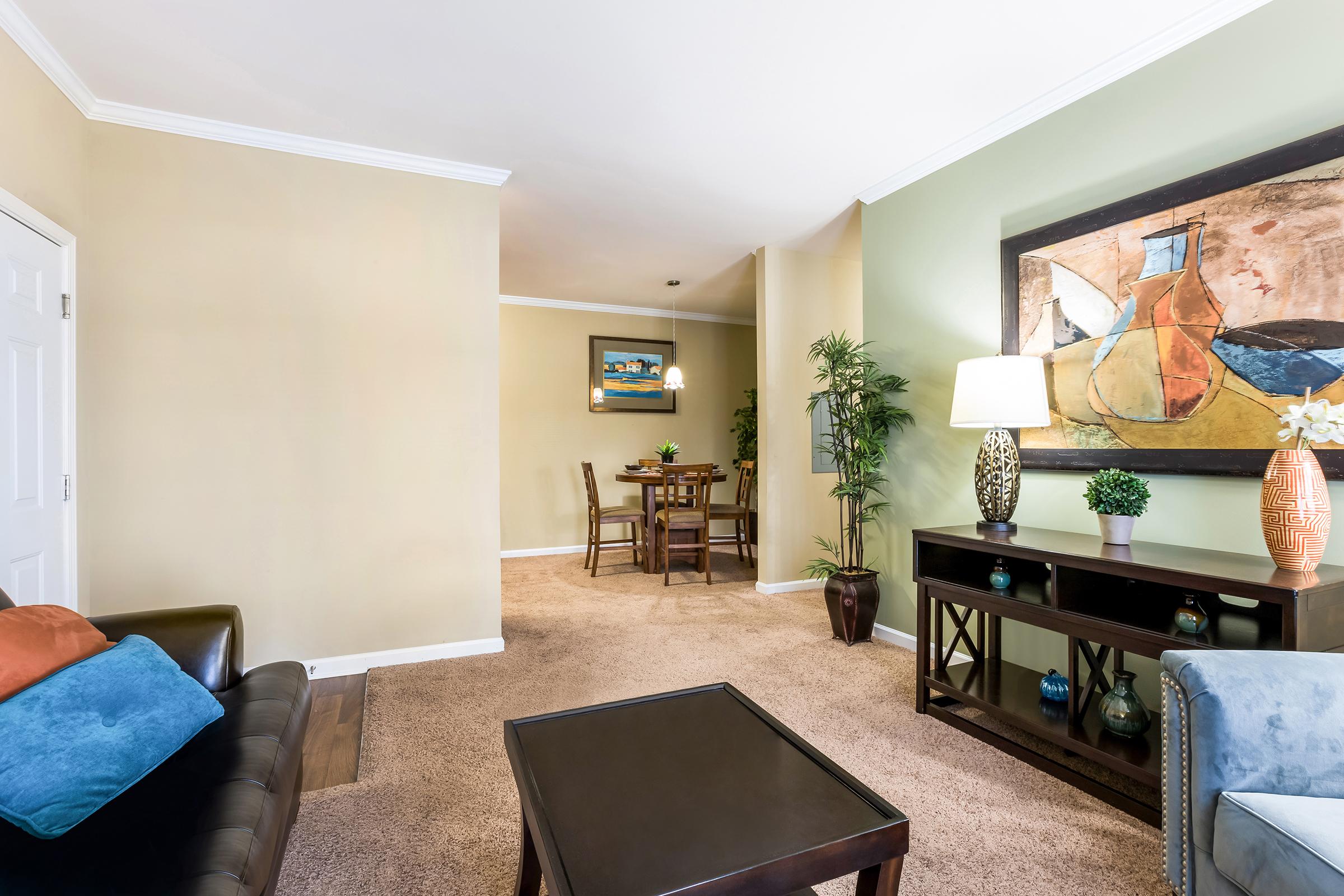 Cozy living room with beige walls and carpeted flooring. A dark couch with blue and orange cushions faces a low coffee table. To the right, a modern painting hangs above a console table featuring a lamp and decorative plants. In the background, a dining area with wooden chairs is visible.