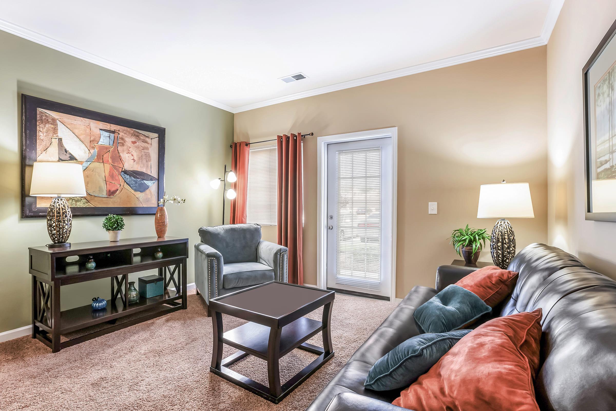 A cozy living room featuring a black leather sofa with colorful pillows, a wooden coffee table, and a console table with decorative items. Two lamps provide soft lighting, and a chair is positioned near a sliding glass door leading to the outside, while a large artwork adorns the wall.