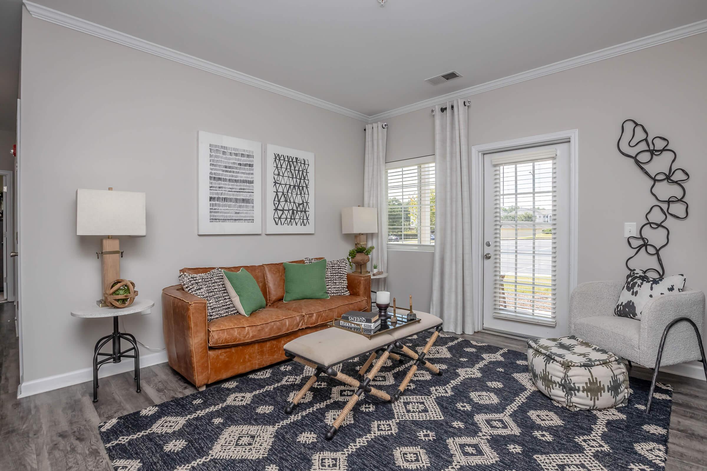 Cozy living room featuring a brown leather sofa with green throw pillows, a round side table with a lamp, and a decorative chair. A patterned rug covers the floor, while large windows with blinds provide natural light. Wall art and modern decor enhance the stylish ambiance of the space.