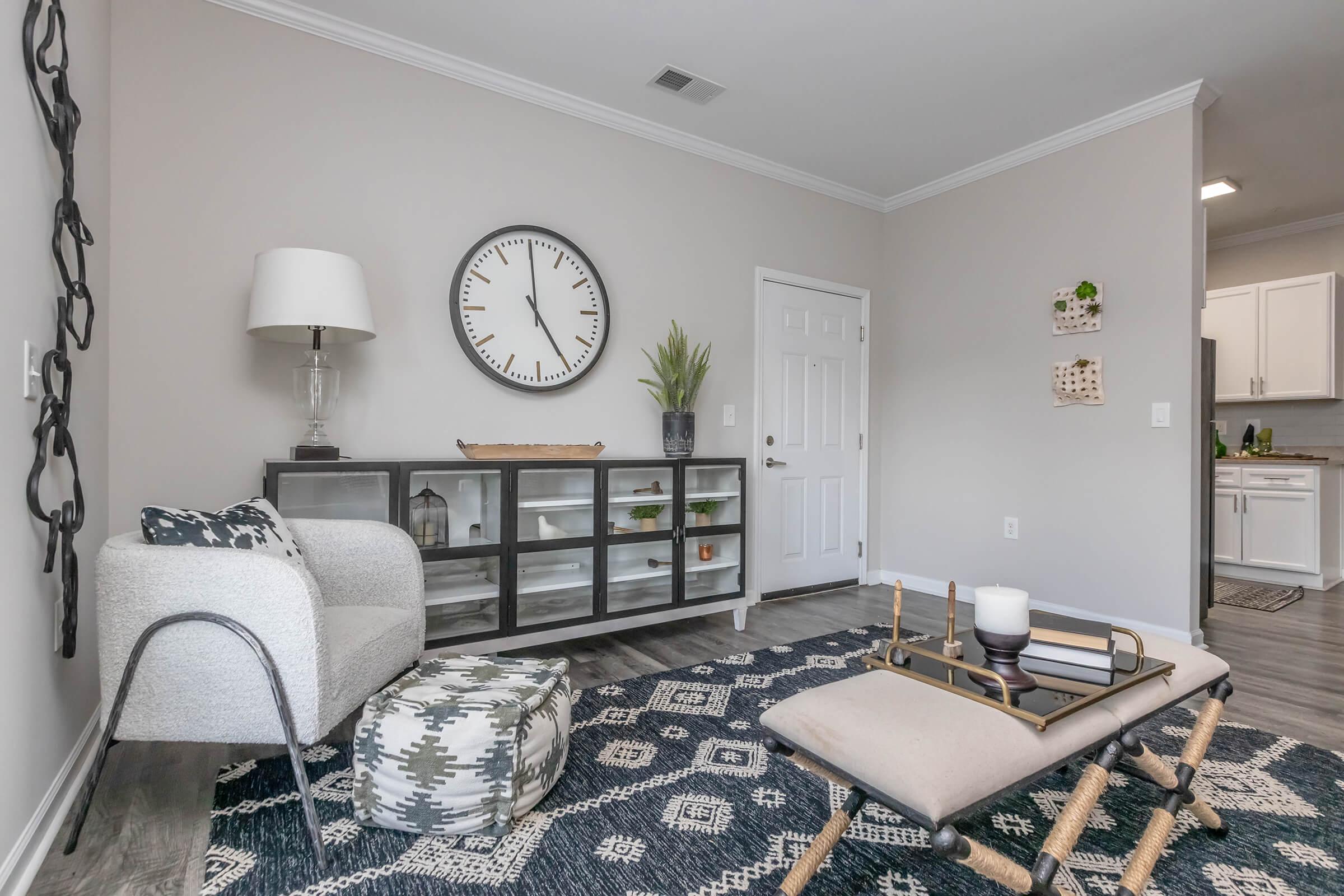 A modern living room featuring a neutral color palette. A stylish armchair sits next to a glass-front cabinet displaying decorative items. A large wall clock is above the cabinet, and a patterned area rug covers the floor. A small plant adds a touch of greenery, with a door leading to an adjoining space in the background.