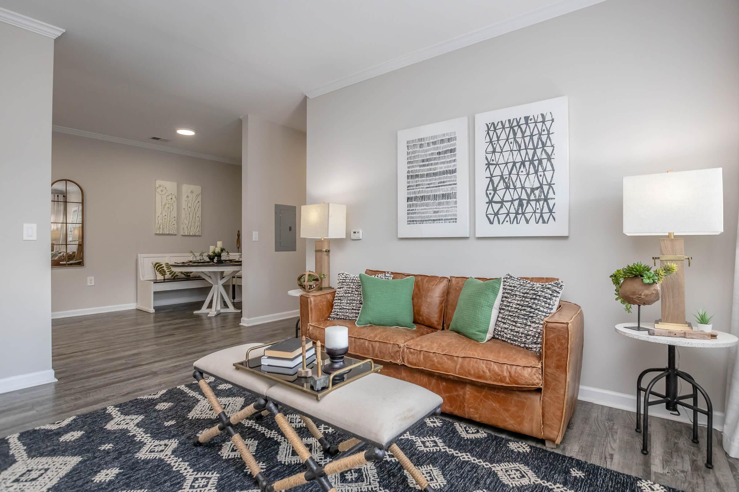 A cozy living room featuring a brown leather sofa adorned with green and patterned pillows. A coffee table with books and decor items sits in front. Two framed artworks hang on the wall, and there are floor lamps on either side of the sofa. The space has modern, neutral tones with a patterned area rug.