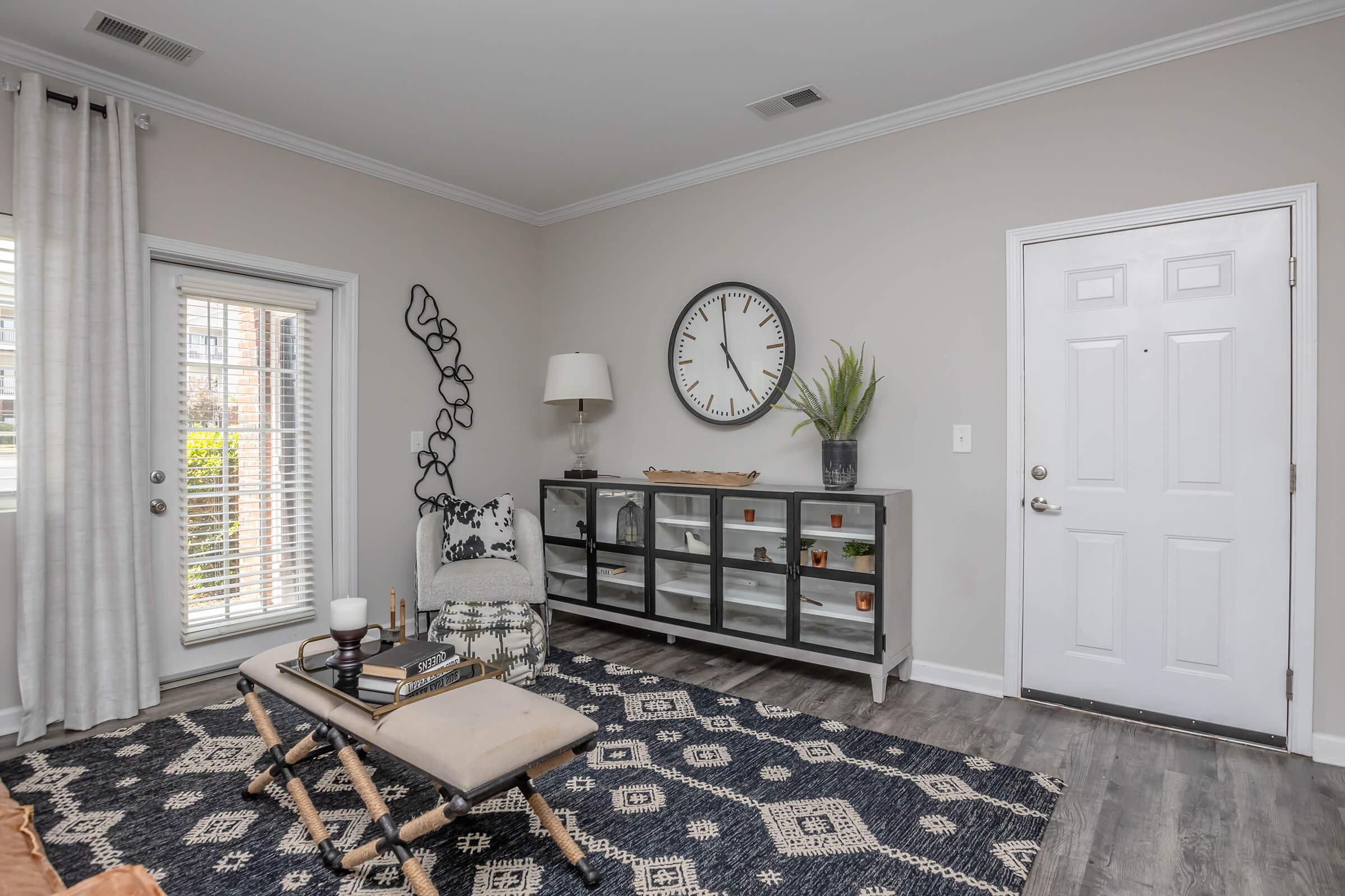 A cozy living room featuring a gray accent wall, a large wall clock, and a modern console table with decorative items. There’s a comfortable seating area with a patterned chair and ottoman, a soft area rug, and a close entrance with a door and window, bringing in natural light.