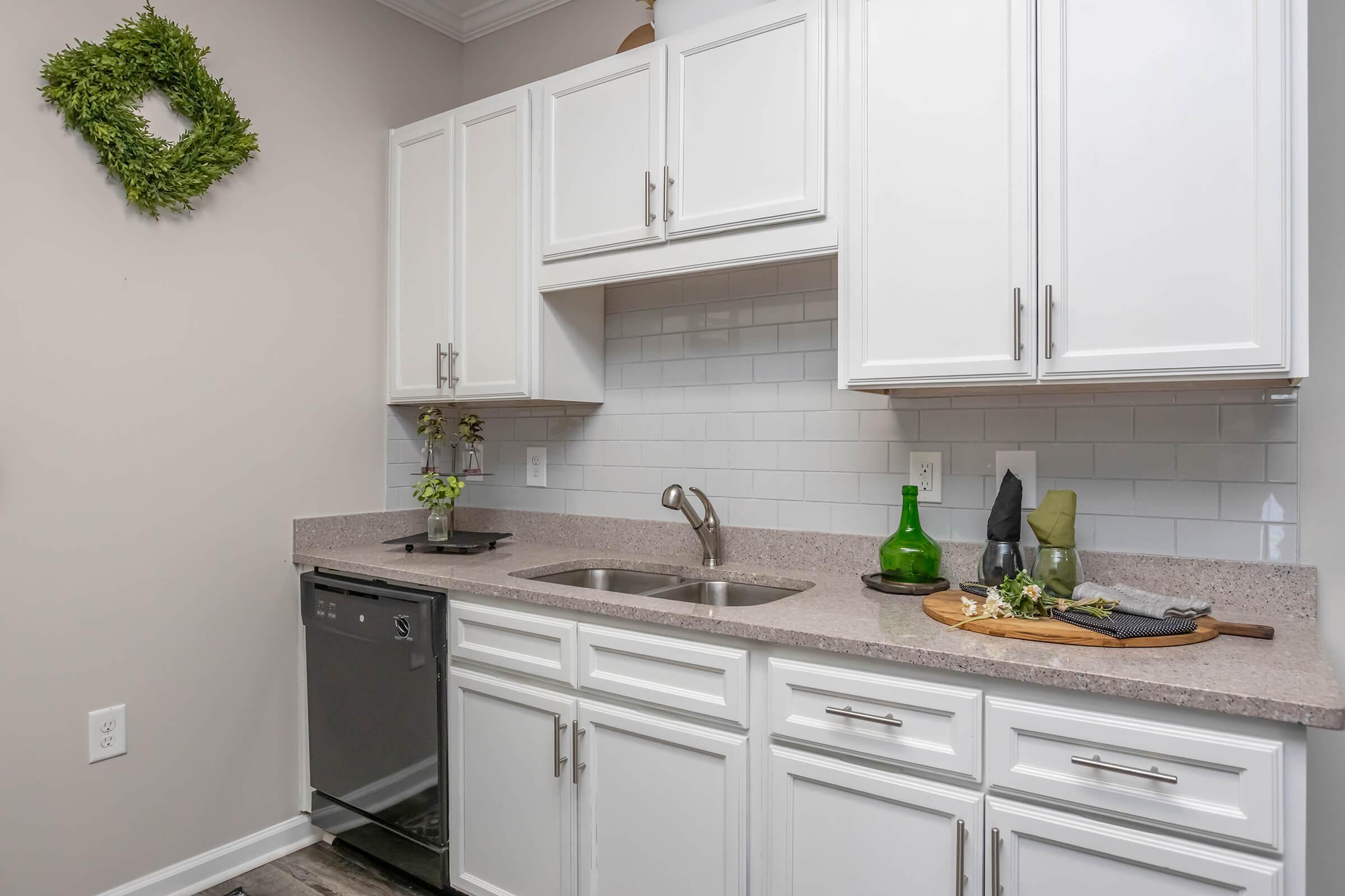 A modern kitchen featuring white cabinetry, a double sink, a stainless steel dishwasher, and a light gray countertop. Decorative elements include a small plant on the counter, a green glass bottle, and a wooden serving tray with utensils. The walls are painted a neutral color, creating a bright and inviting space.