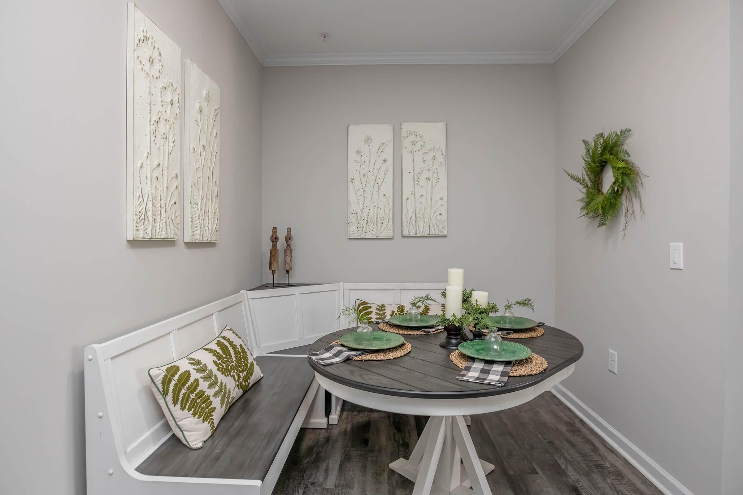 A cozy dining nook featuring a round table with green plates and woven placemats. The bench is adorned with a leafy cushion. Three textured wall art pieces and a small greenery wreath hang above, adding a touch of nature, while soft lighting enhances the inviting atmosphere.