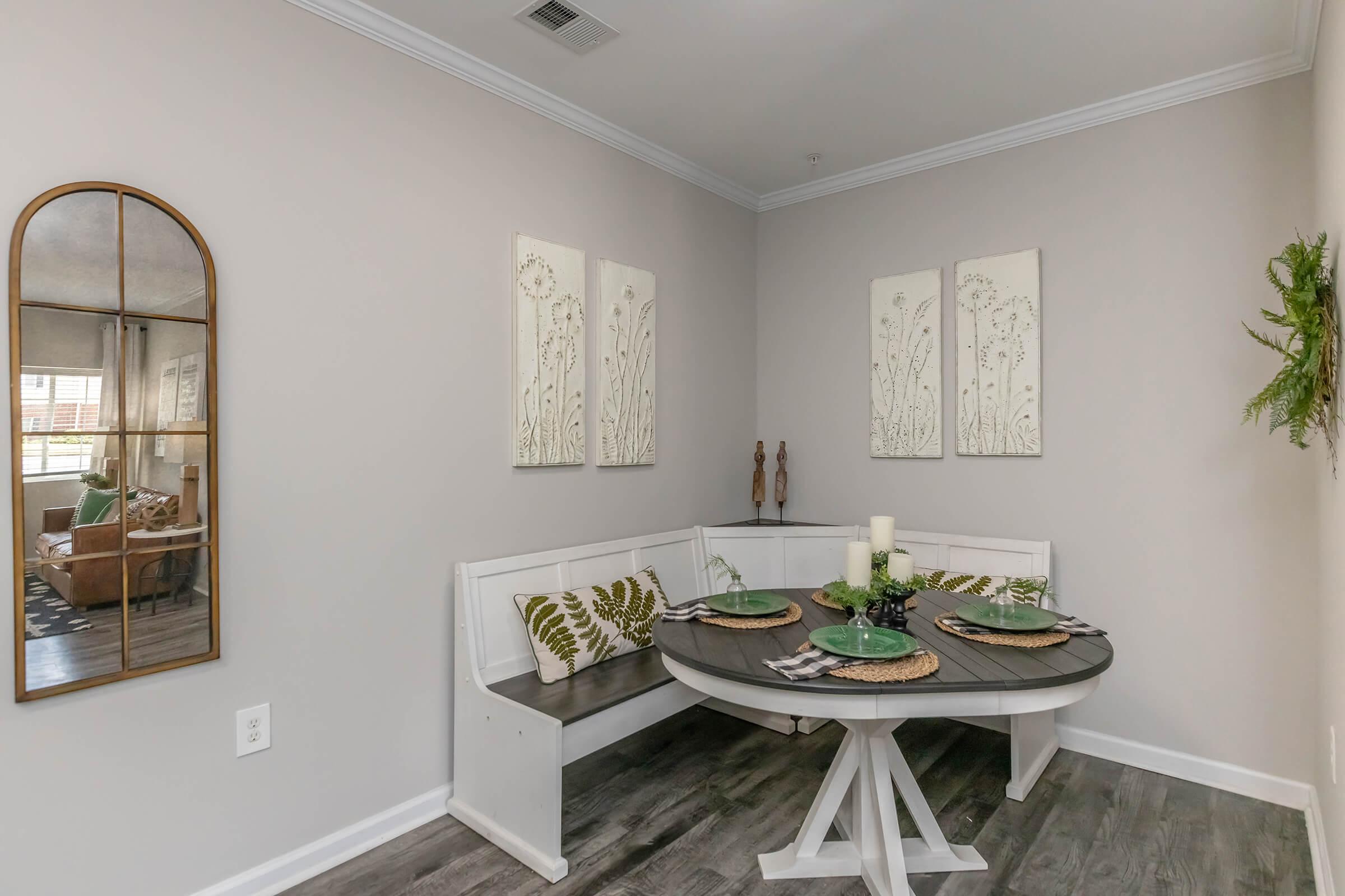 A cozy dining area featuring a round table with decorative plates and utensils, a built-in bench with cushions, and wall art of floral designs. A mirror is hung on one wall, reflecting the space, while decorative green accents add a natural touch to the neutral color scheme.