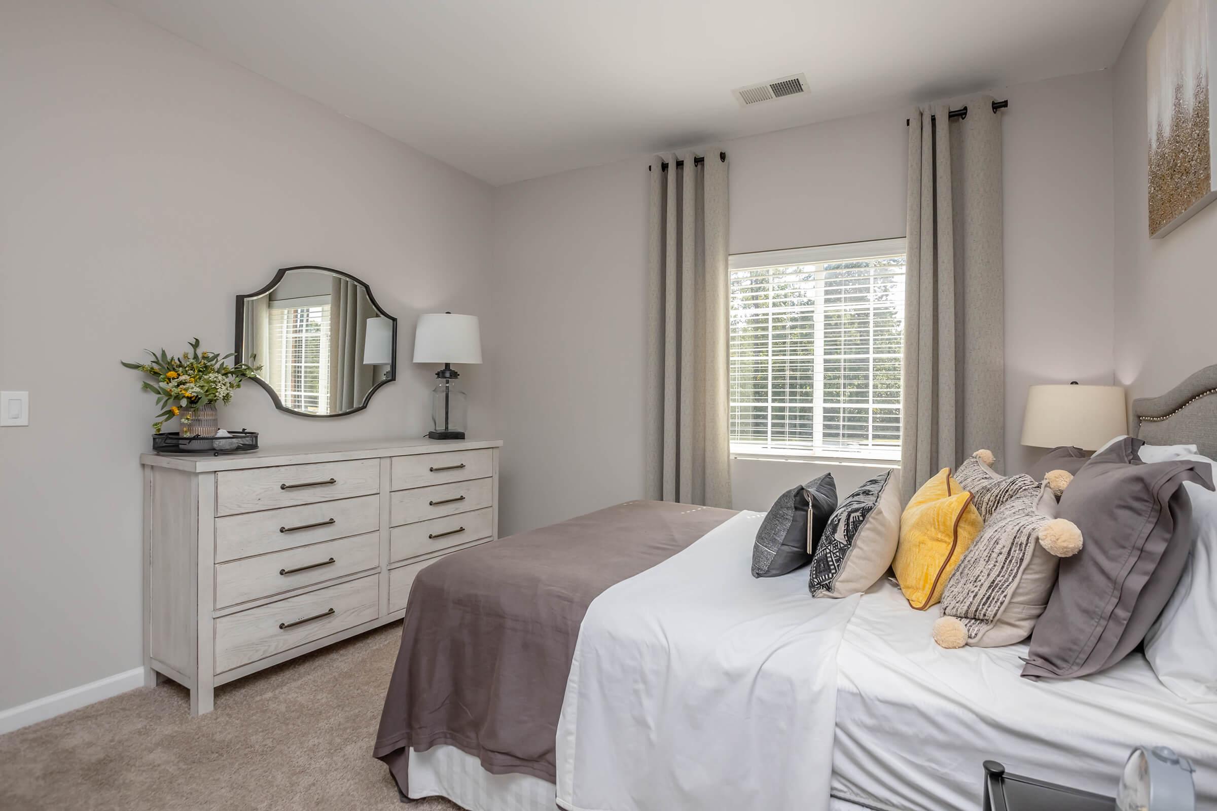 A cozy bedroom featuring a neatly made bed with decorative pillows, a light-colored dresser, a lamp, and a mirror. Natural light filters through a window adorned with gray curtains. A small vase with flowers adds a touch of freshness to the room’s calming atmosphere.