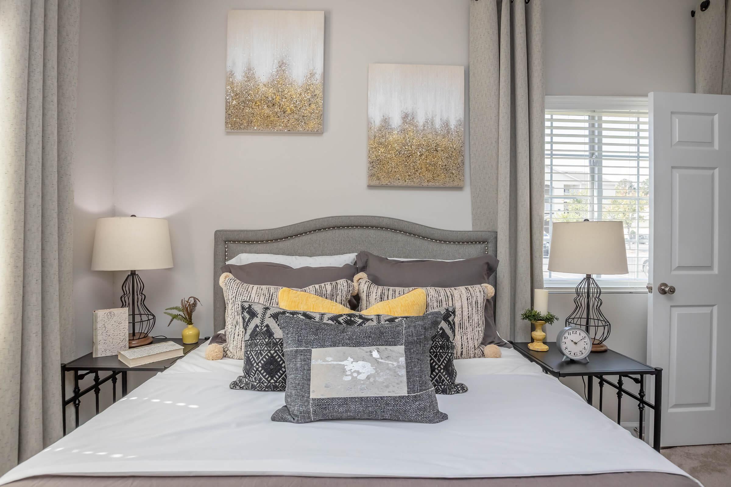 A stylish bedroom featuring a neatly made bed with multiple pillows, gray and yellow accents, and two bedside tables with lamps. Above the bed, there are two abstract art pieces. The room is illuminated by natural light from a window with blinds, and there's a small plant and clock on the nightstand.