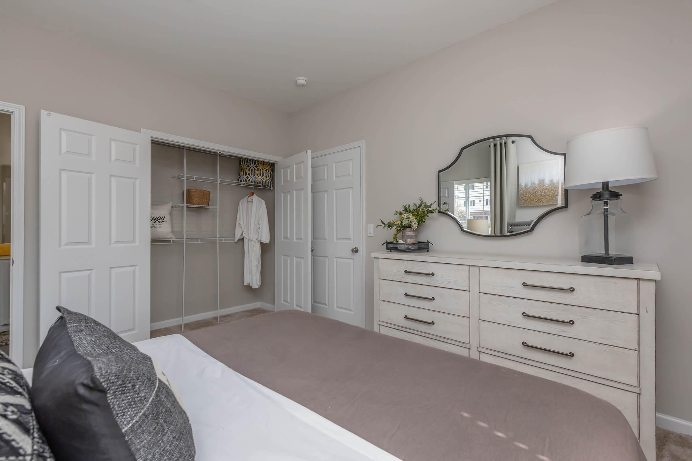 A well-decorated bedroom featuring a comfortable bed with gray bedding, a light wooden dresser with multiple drawers, and a round mirror. There is a closet with open doors showing a robe and towels, and a stylish lamp on the dresser. Soft, neutral wall colors create a calming atmosphere.