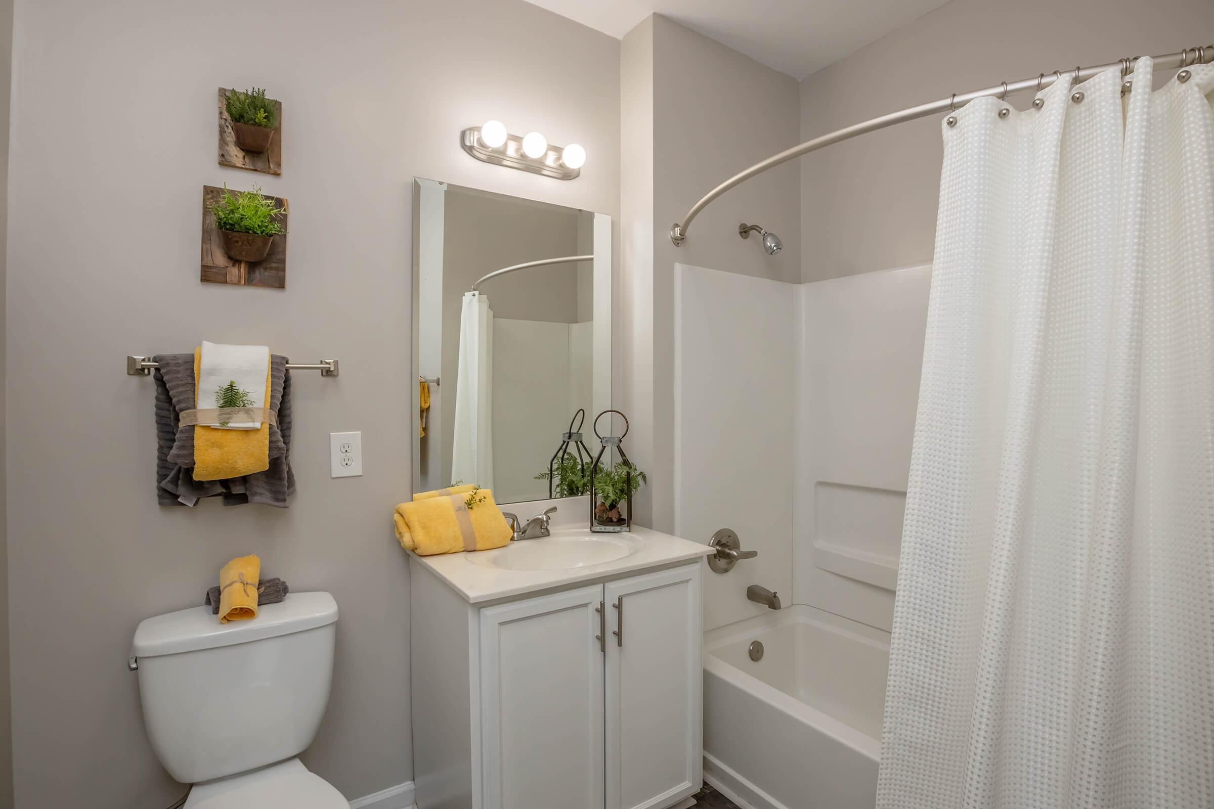 A modern bathroom featuring a white bathtub and shower, a white vanity with a mirror, and a toilet. The walls are painted in a light gray color, and decorative elements include potted plants on wall shelves and neatly arranged towels on a rack. Bright yellow accents add a pop of color.
