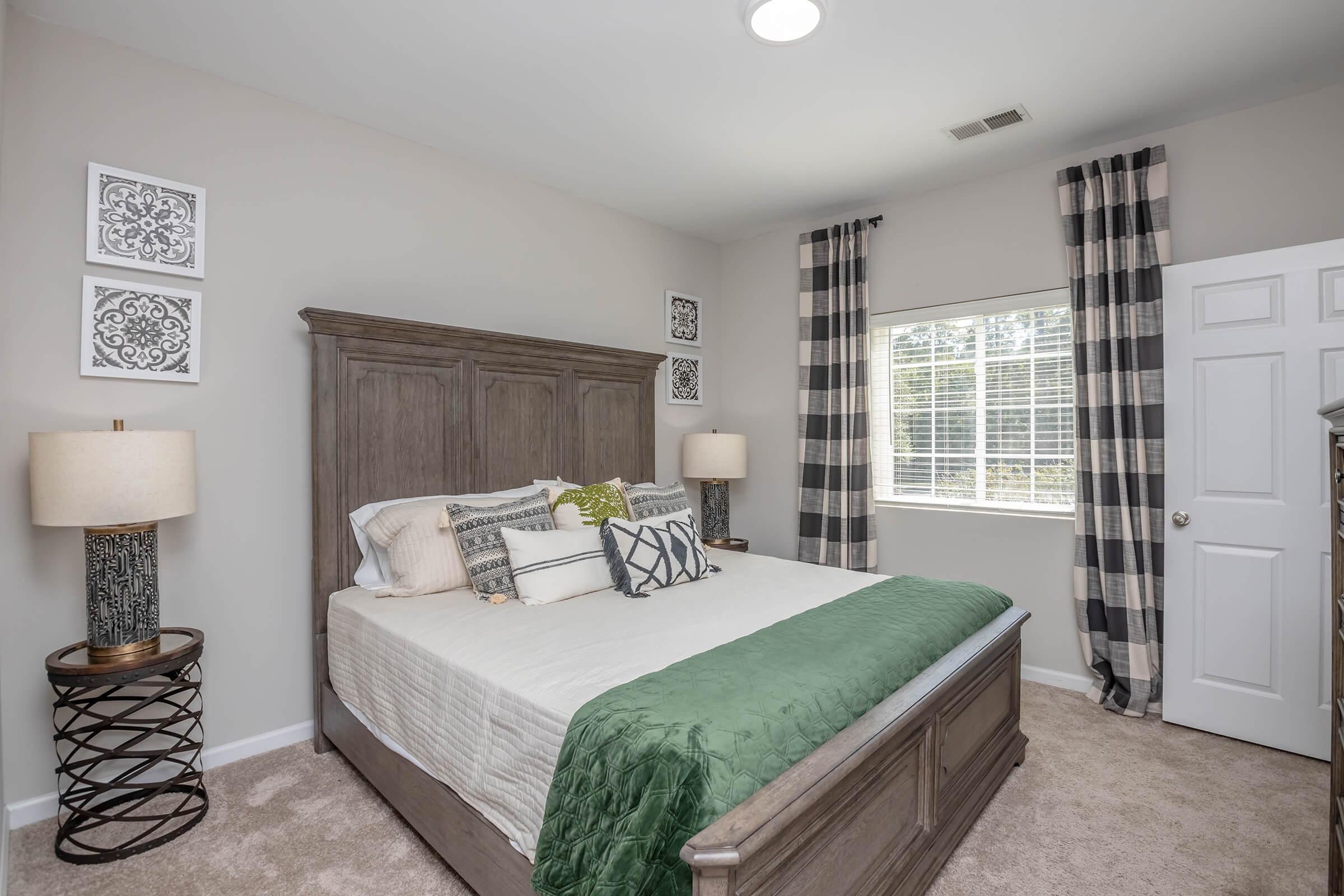 A cozy bedroom featuring a queen-sized bed with a green throw blanket, decorative pillows, and a wooden headboard. There are two lamps on nightstands, patterned curtains, and framed artwork on the walls. A window allows natural light to brighten the room, which has neutral-colored walls and carpet.