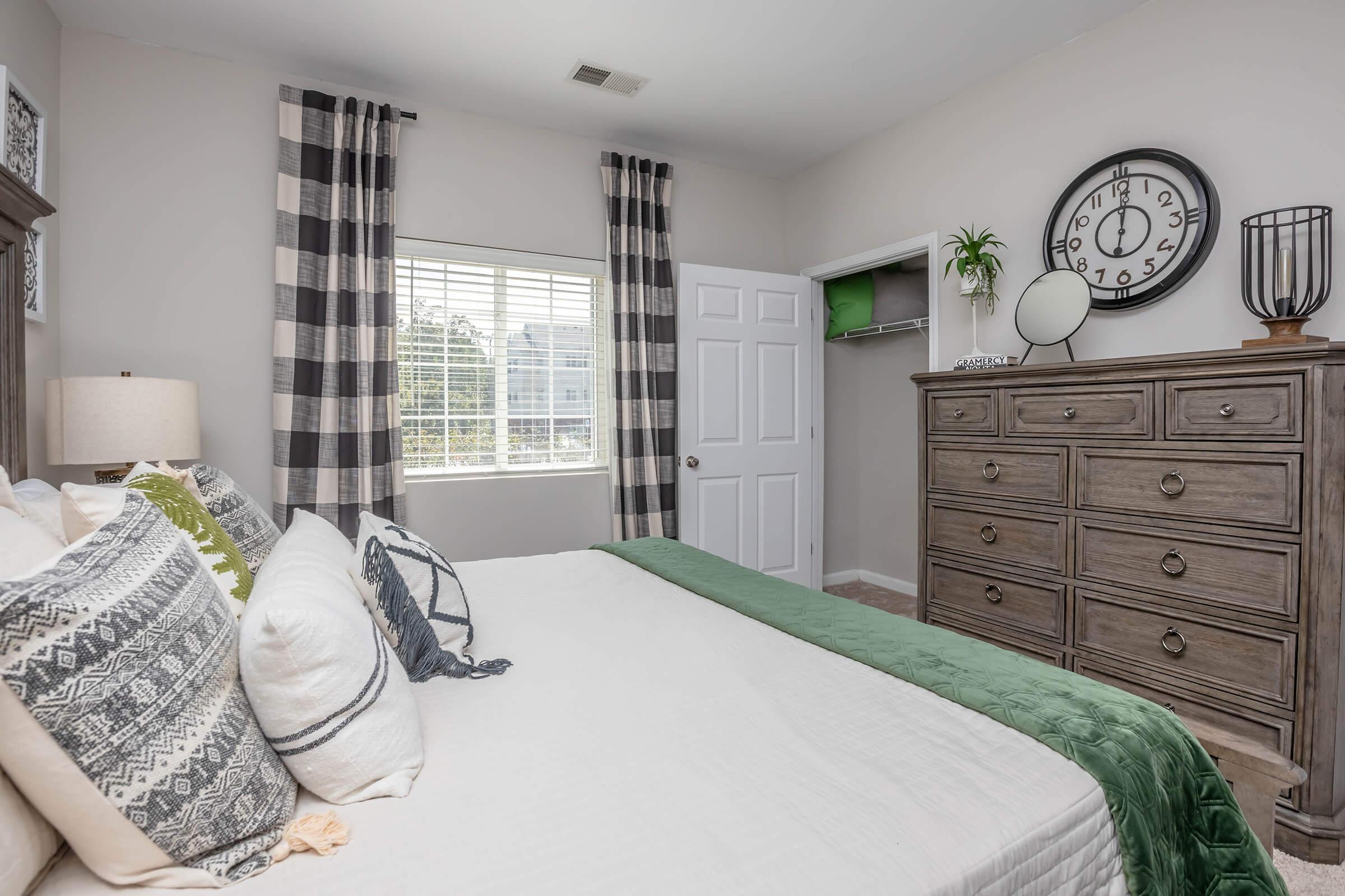 A well-decorated bedroom featuring a large bed with decorative pillows and a green throw blanket. There's a dresser with decorative items, a wall clock, and a window with sheer curtains allowing natural light. The room has neutral-colored walls and an organized closet space.