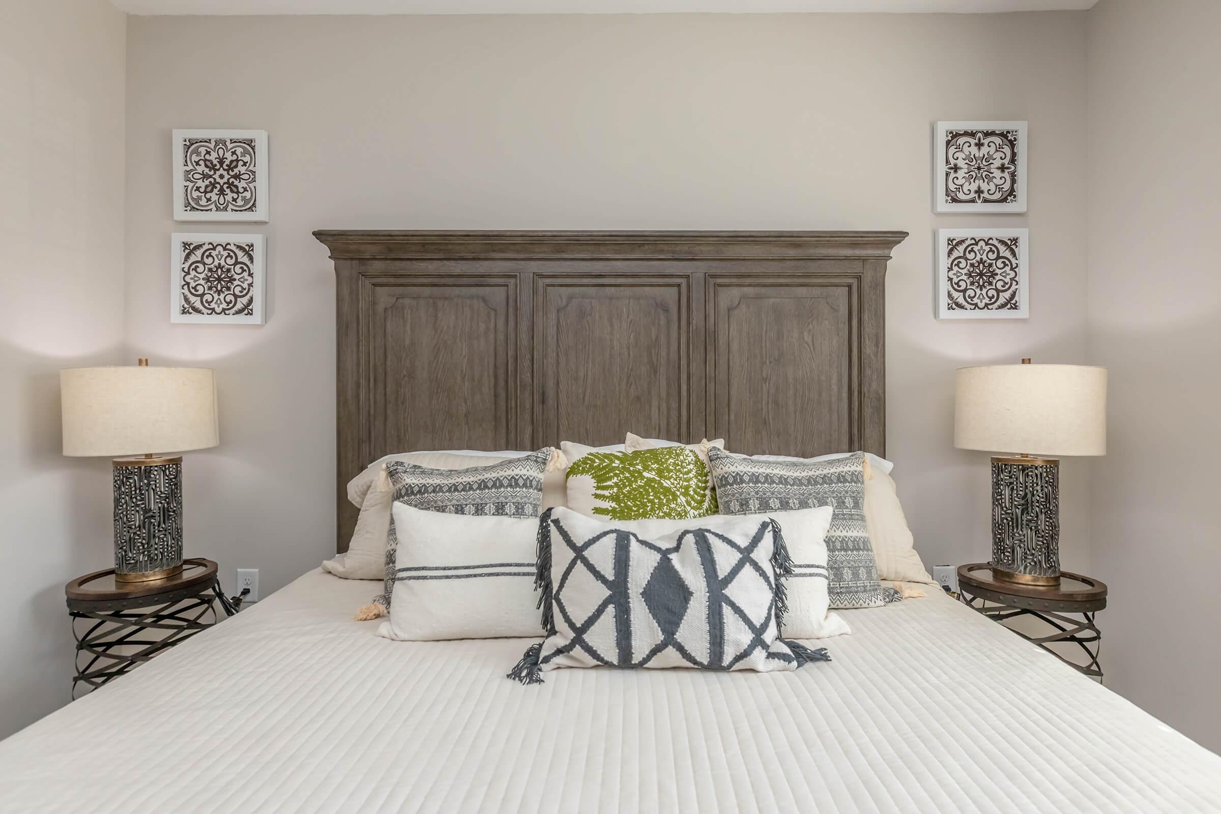 A cozy bedroom featuring a wooden headboard with decorative elements, a neatly made bed adorned with various textured pillows, and two stylish lamps on nightstands. Four framed artworks are displayed on the wall above the bed, contributing to a warm and inviting atmosphere.