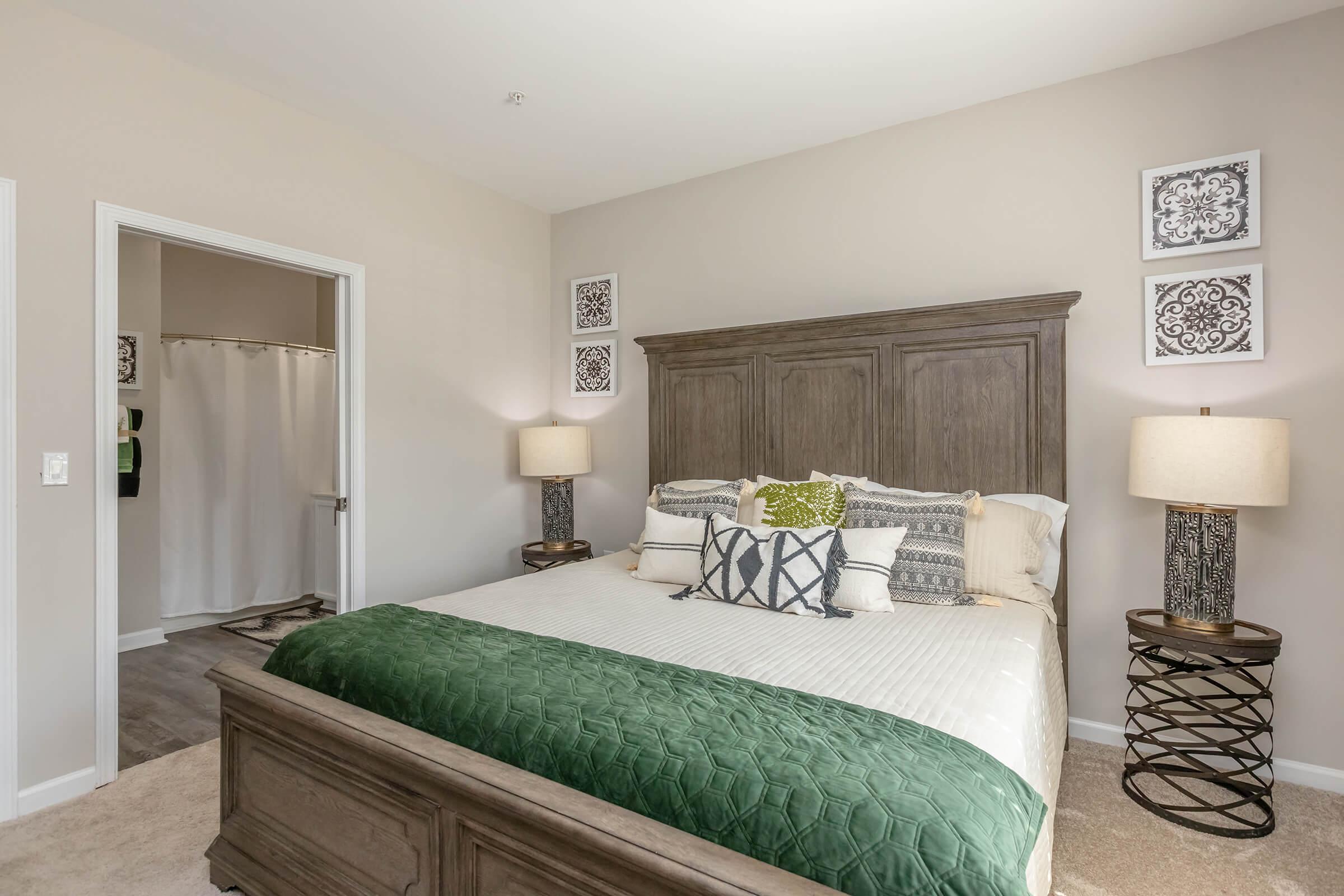 A cozy bedroom featuring a large bed with a green and white quilt, decorative pillows, and two bedside lamps on woven side tables. Light-colored walls and framed art add elegance, while a bathroom can be seen in the background through an open door.