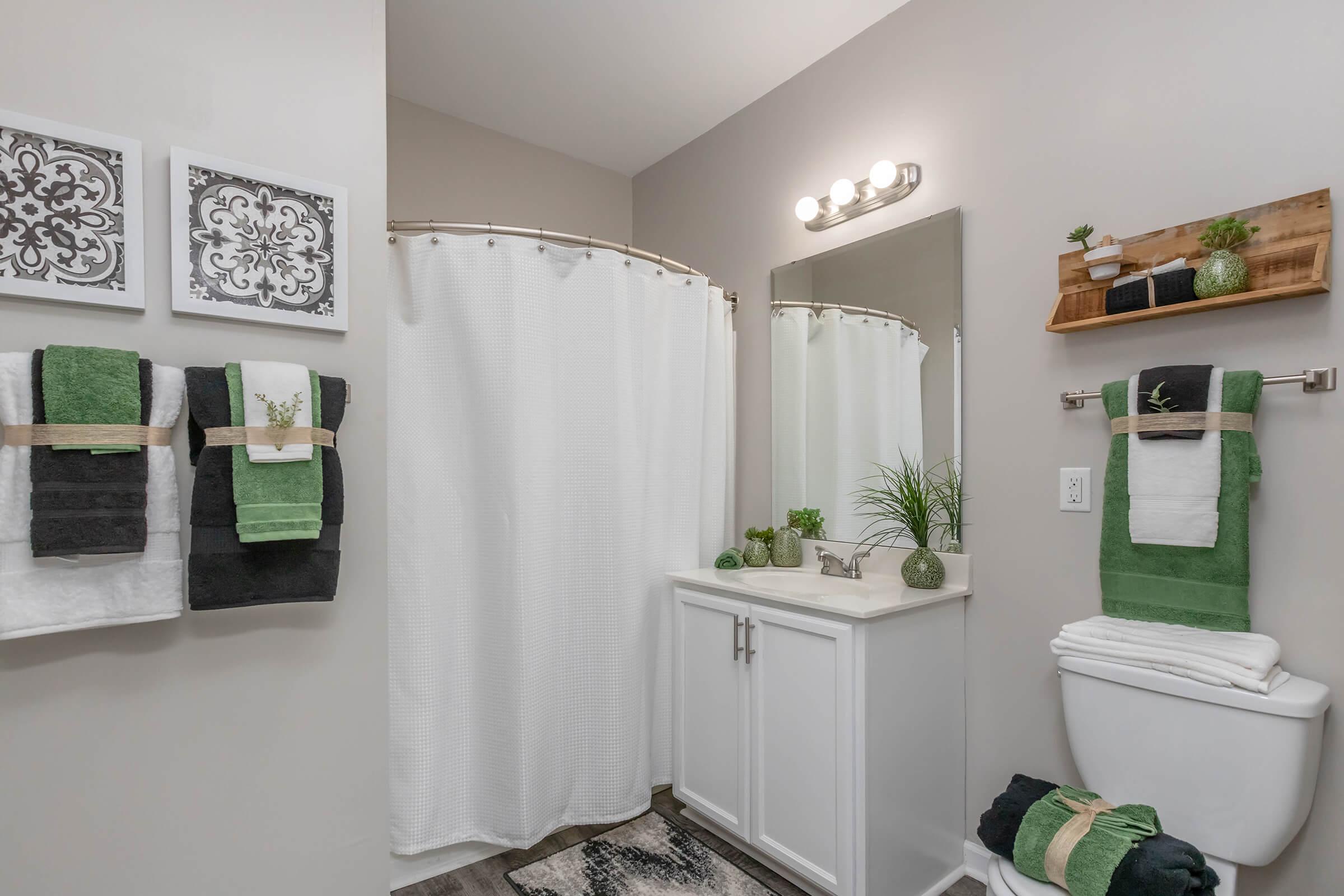 A modern bathroom featuring a white vanity with a mirror, a shower curtain, and neatly arranged towels in green and black. Decorative elements include artwork on the walls, a small shelf with plants, and neatly stacked white towels on the toilet. The overall decor is clean and inviting.