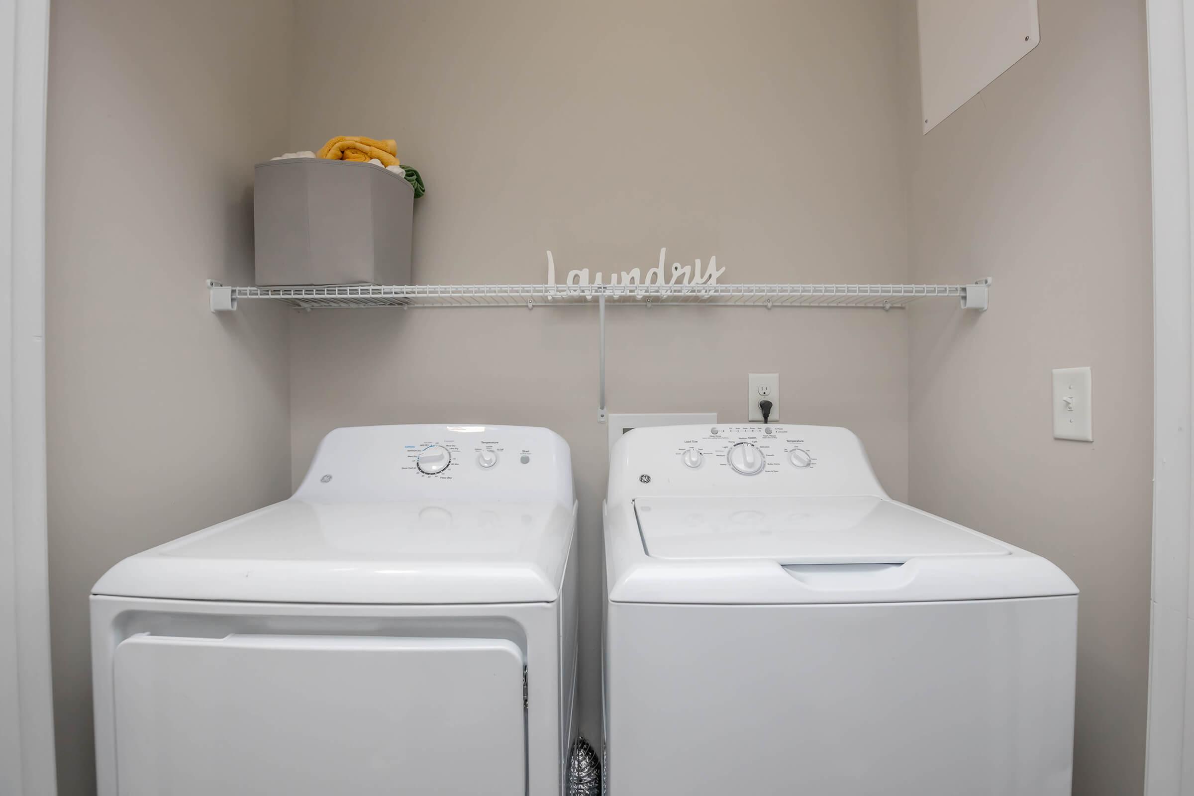 A clean and organized laundry room featuring a white washer and dryer side by side. Above them, a metal wire shelf holds a gray storage box and a folded yellow towel. A decorative sign reading "Laundry" is displayed on the shelf, enhancing the room's aesthetic. The walls are a soft beige color.