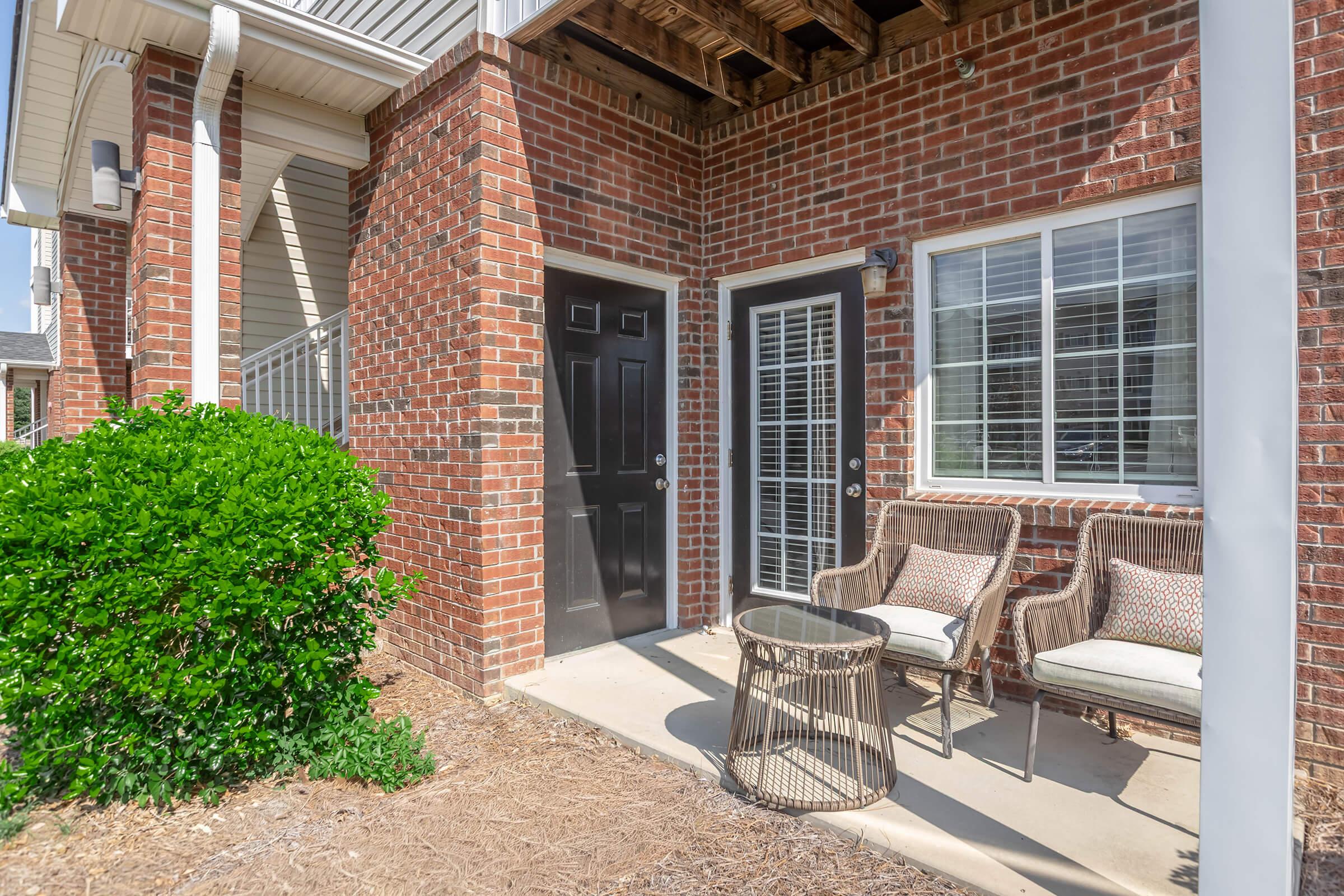 Front entry area of an apartment featuring two black doors, a large window with multiple panes, and a small round table with two wicker chairs. Lush green shrubbery is present in front of the brick exterior, which has a mix of red and tan bricks, and a covered porch area above.