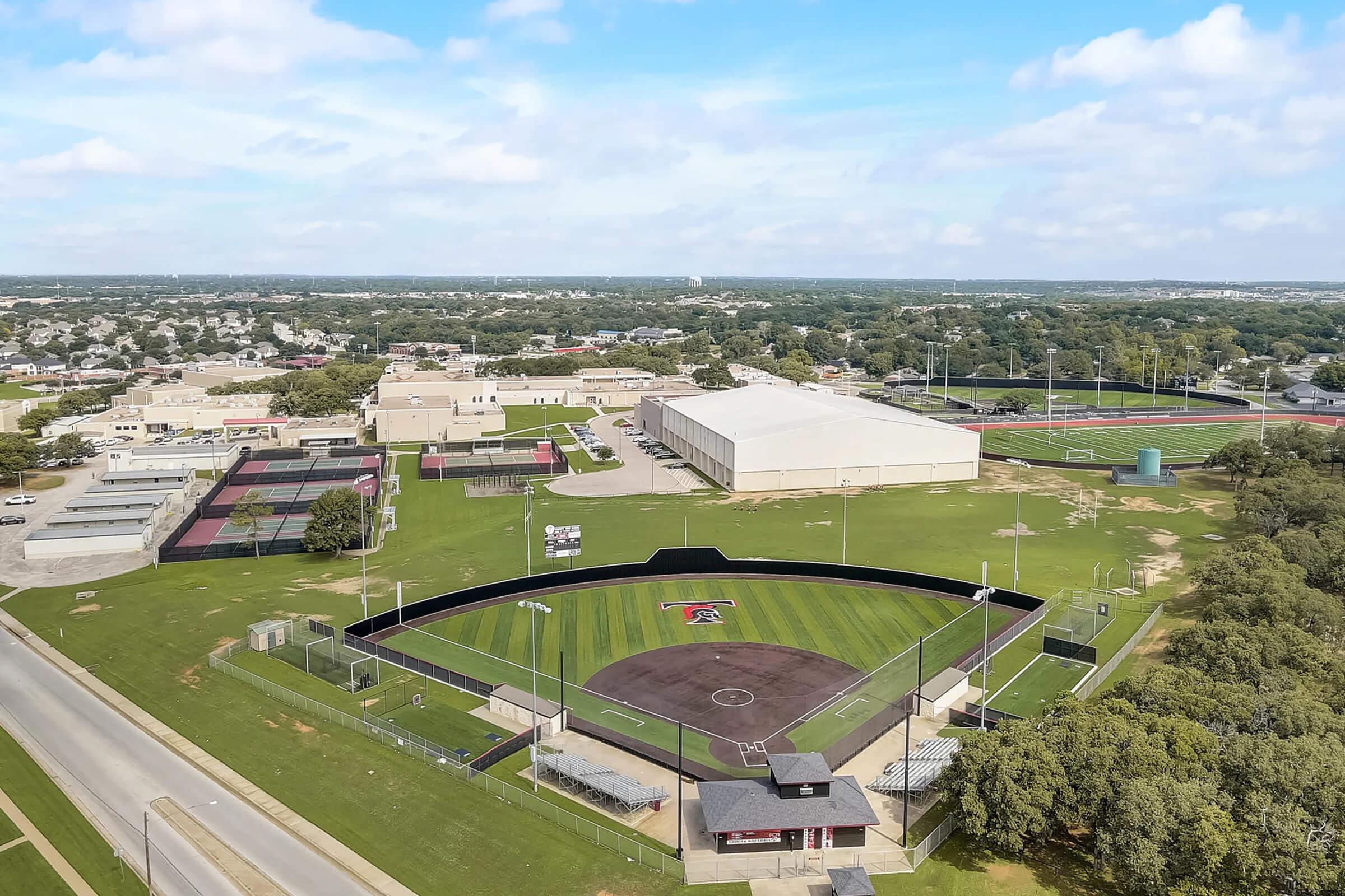 Aerial view of a sports complex featuring a baseball field with a black outfield fence, surrounded by grassy areas and bleachers. Nearby buildings include a gymnasium and athletic facilities. The landscape is dotted with trees and the sky is partly cloudy, indicating a nice day for outdoor activities.