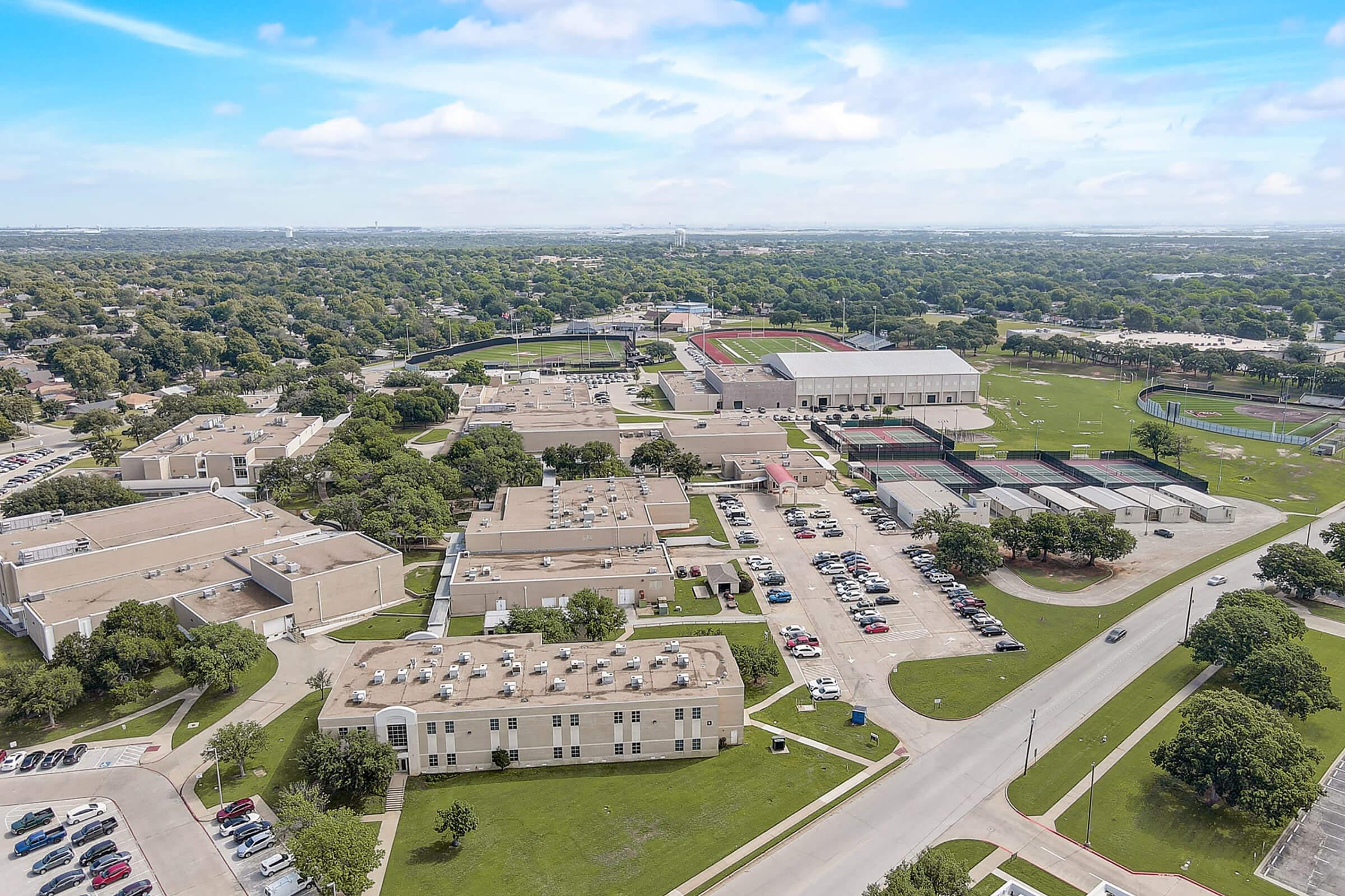 Aerial view of a large educational campus featuring multiple buildings, sports fields, and parking lots, surrounded by green trees and open spaces under a partly cloudy sky. The layout shows a combination of residential areas and athletic facilities.