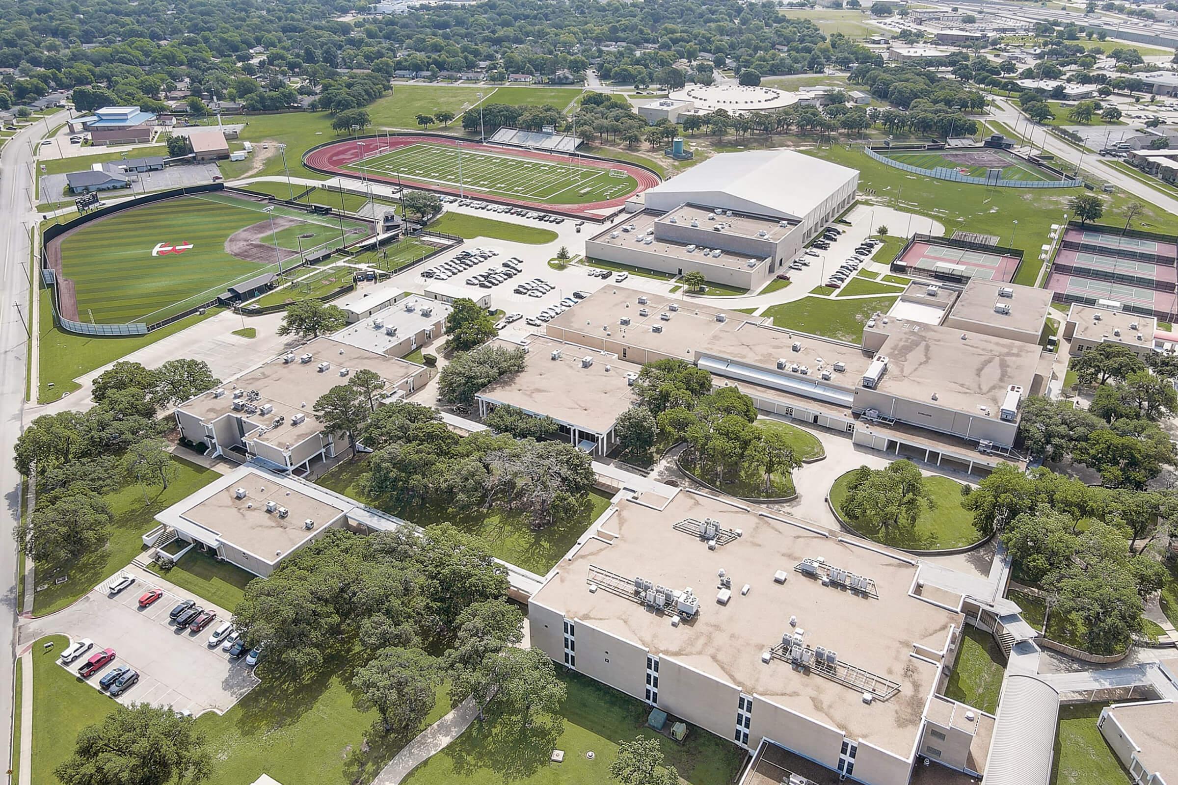 Aerial view of a school campus featuring multiple buildings, green fields, a track and football field, and tennis courts. The area is surrounded by trees and has a parking lot with several vehicles. The layout shows a well-maintained educational facility with sports amenities.