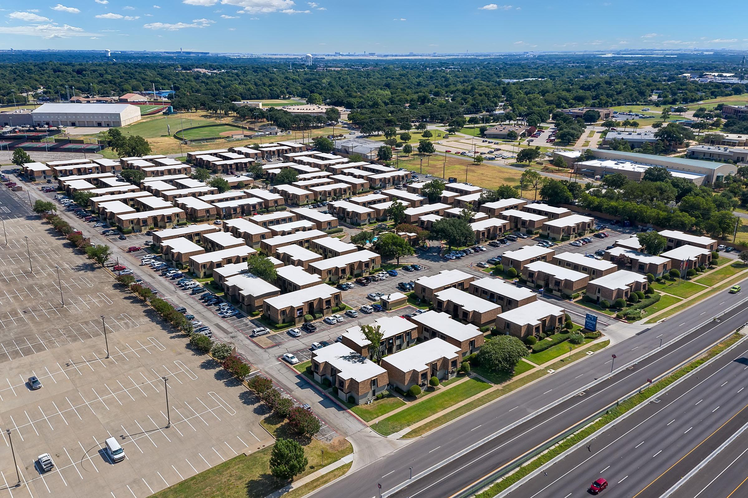 Aerial view of a residential complex featuring multiple buildings with flat roofs, surrounded by green spaces and parking lots. In the background, there are more buildings and a vast landscape under a clear blue sky. The scene captures a suburban area with organized rows of homes.