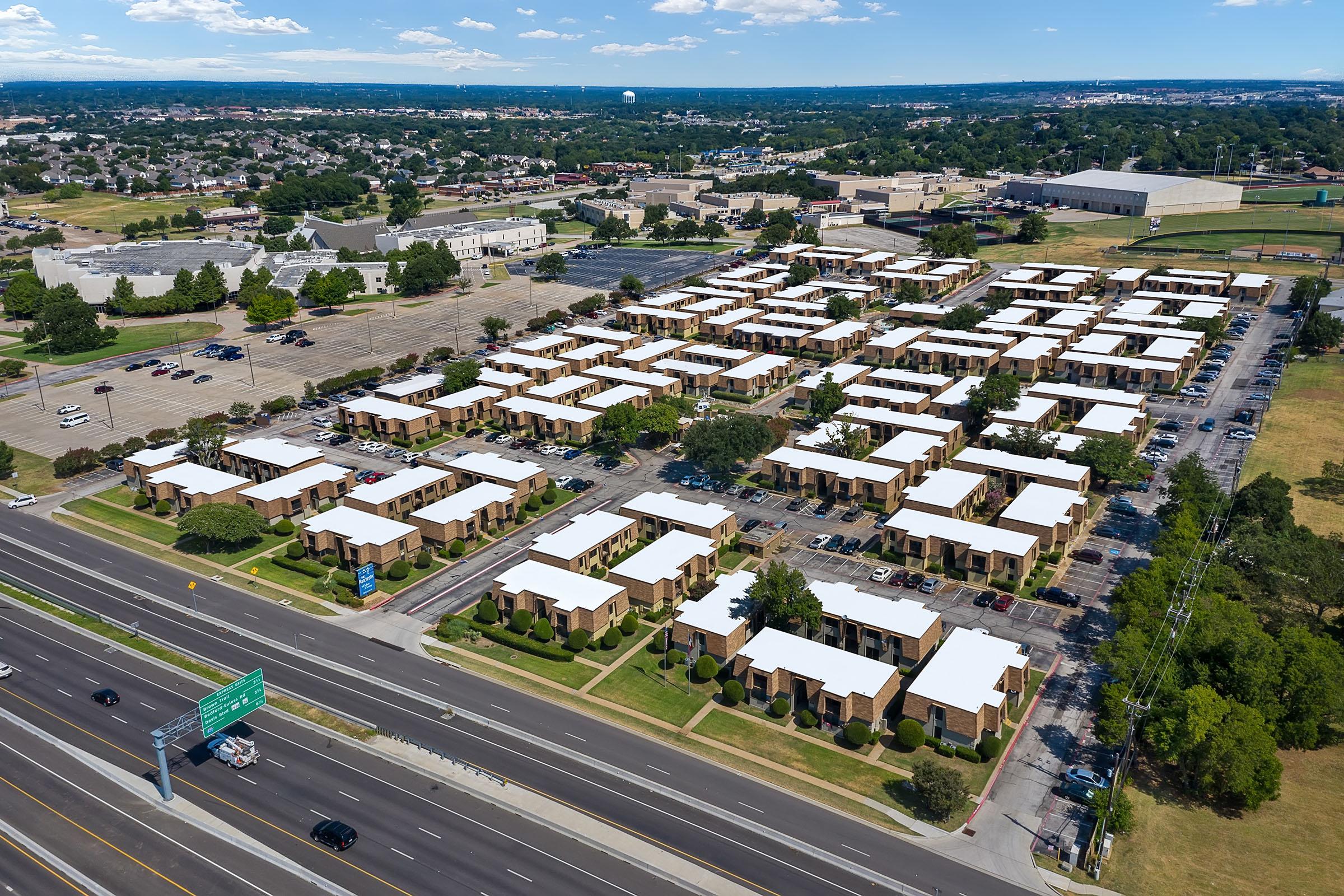 Aerial view of a residential complex featuring multiple single-story buildings with white roofs, surrounded by green grass and trees. Nearby, there are parking spaces and a highway running along the bottom of the image. In the background, additional commercial and residential structures are visible.