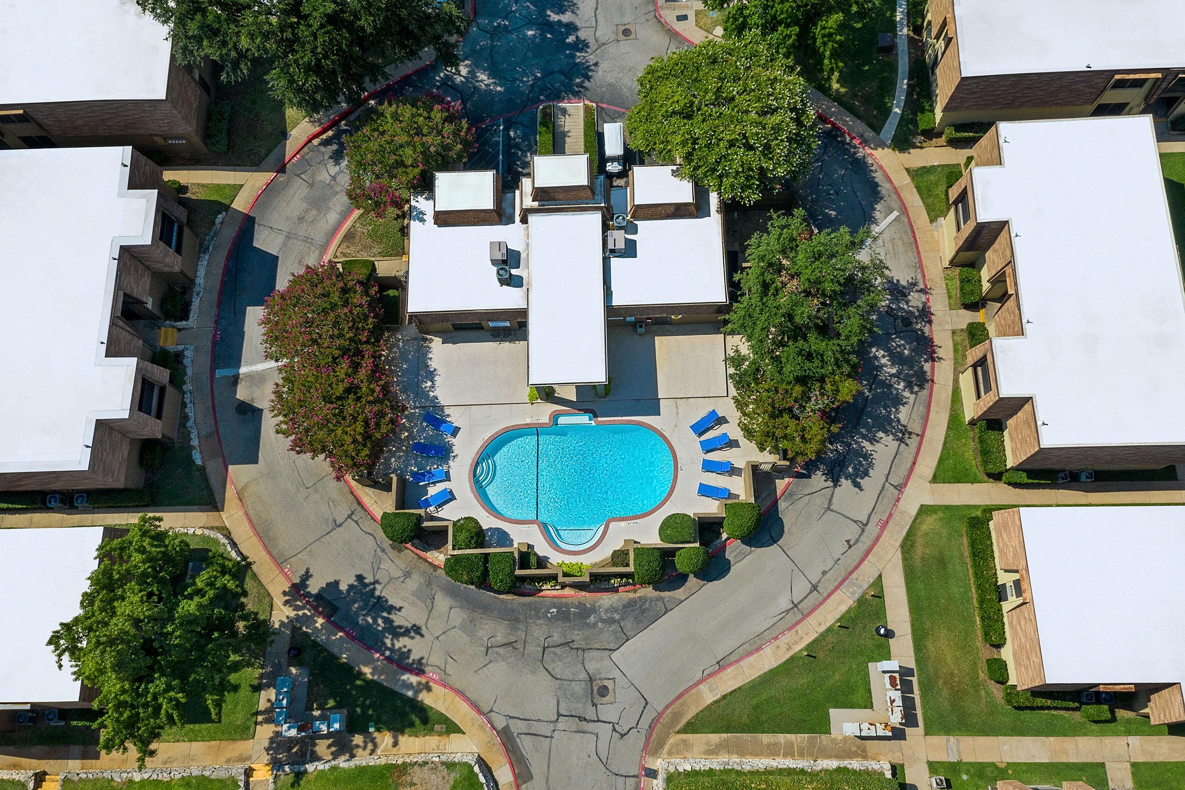 Aerial view of a circular apartment community surrounding a blue swimming pool. The pool area features lounge chairs and is bordered by lush greenery. White-roofed buildings provide a contrast to the vibrant blue of the pool and the manicured lawns. The layout showcases a well-maintained, inviting residential atmosphere.