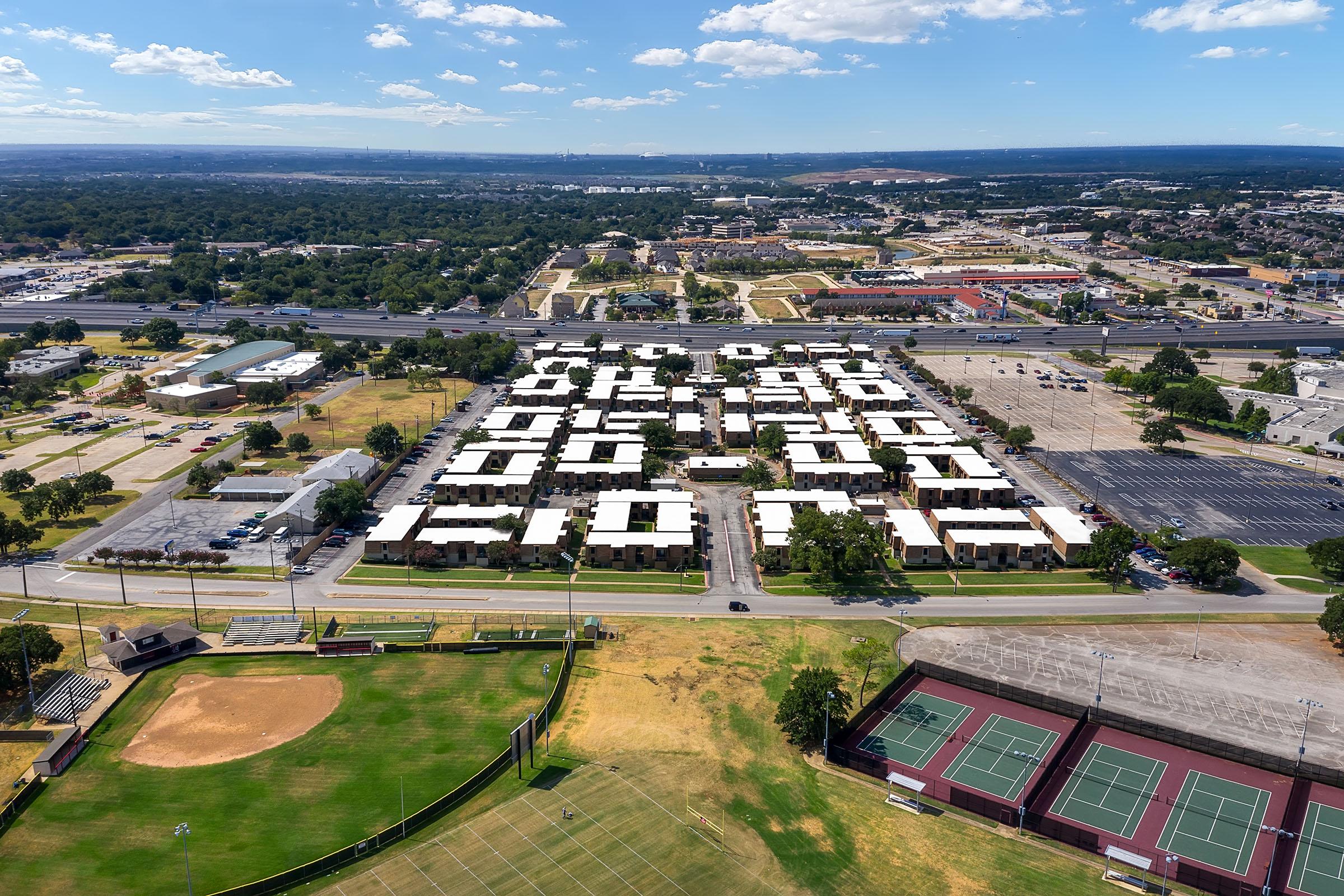 Aerial view of a large complex featuring buildings arranged to form numbers, surrounded by open fields, a baseball diamond, and tennis courts. The landscape includes distant greenery and urban developments, with clear blue skies and scattered clouds.