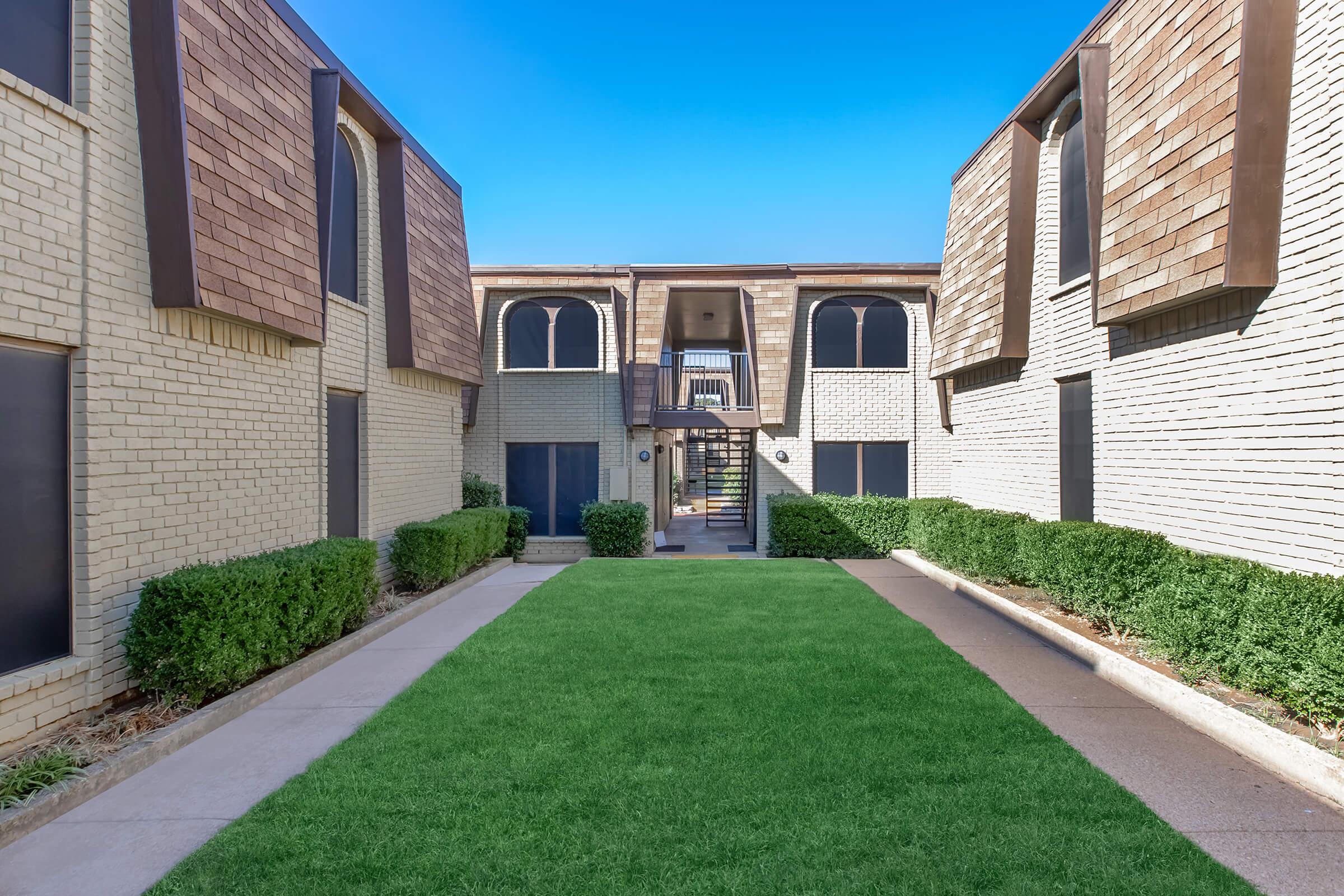 A well-maintained courtyard of an apartment community featuring lush green grass, lined with neatly trimmed shrubs. The architecture includes two buildings with a blend of brick and wooden siding, and a clear blue sky overhead. Pathways lead to the entrance, creating a welcoming atmosphere.