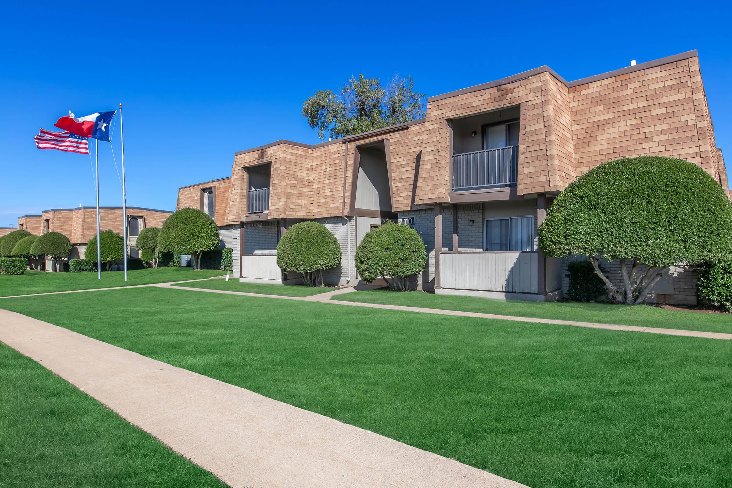 Two multi-story brick apartment buildings with balconies, surrounded by neatly trimmed bushes and vibrant green grass. In front, there are two flags on flagpoles: the American flag and a Texas flag. The sky is clear and blue, creating a bright atmosphere.