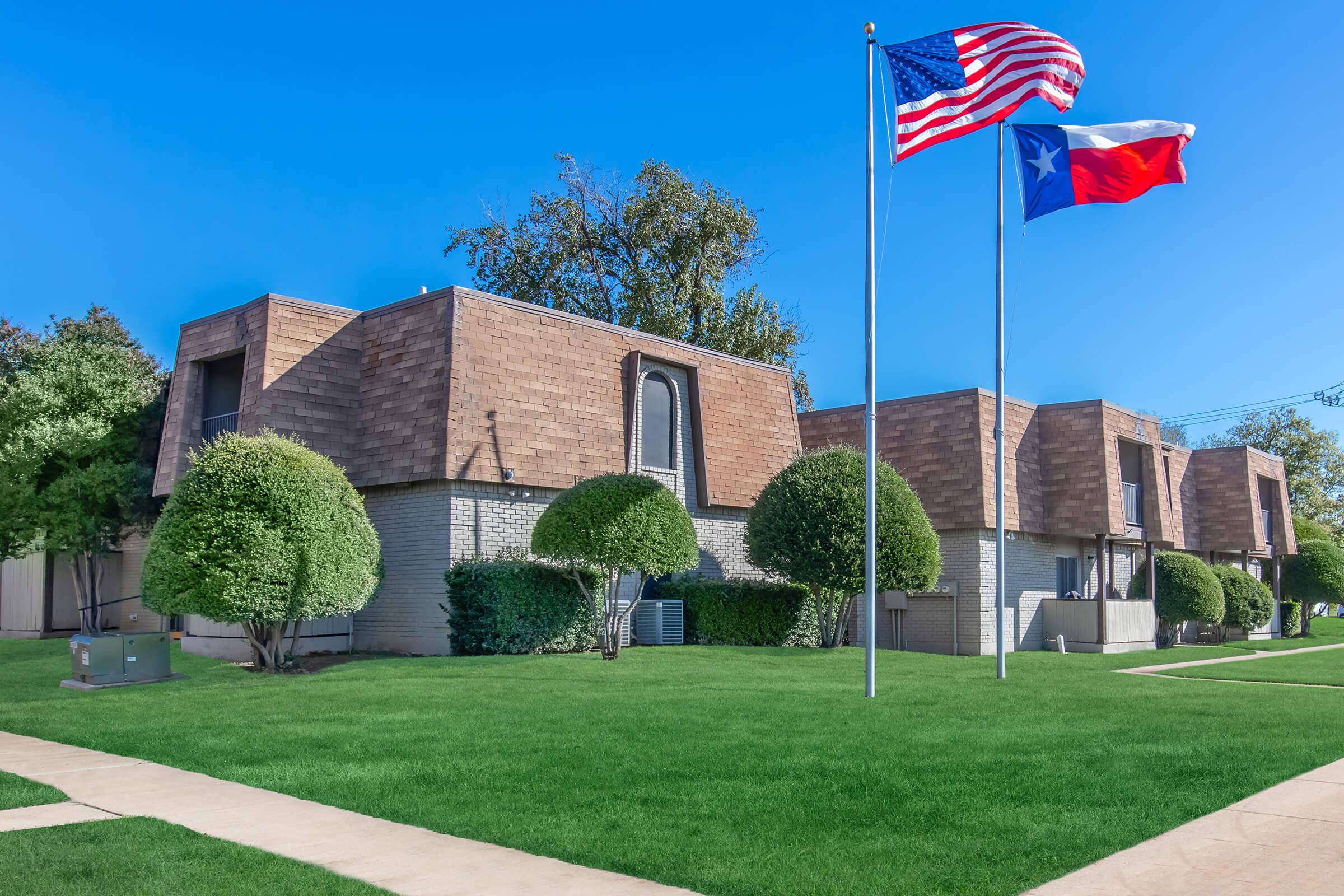 A grassy area in front of a residential complex featuring three buildings with distinctive sloped roofs. Two flags, the United States flag and the Texas flag, are displayed on poles in the foreground, surrounded by neatly trimmed bushes and trees under a clear blue sky.