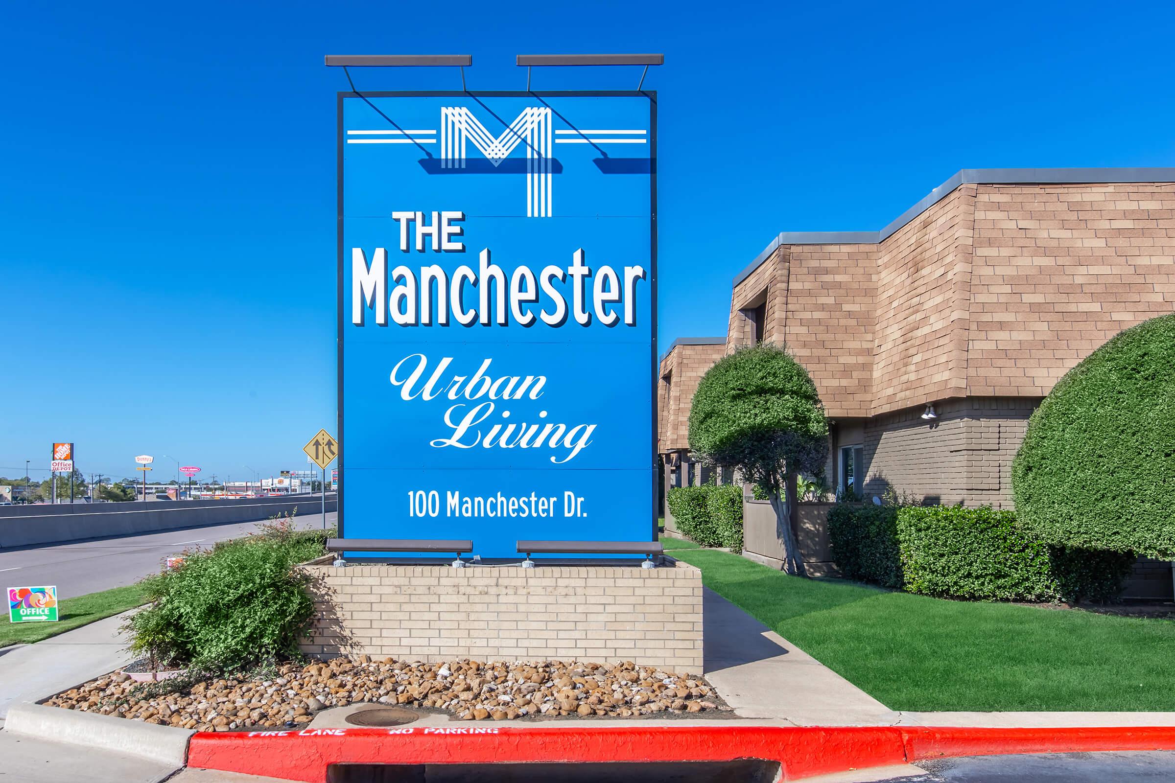 Blue sign for "The Manchester Urban Living" with a large letter "M" at the top, located at 100 Manchester Dr. The sign is positioned near a well-maintained landscaped area with green grass and round shrubs, alongside a driveway marked with red curbing.