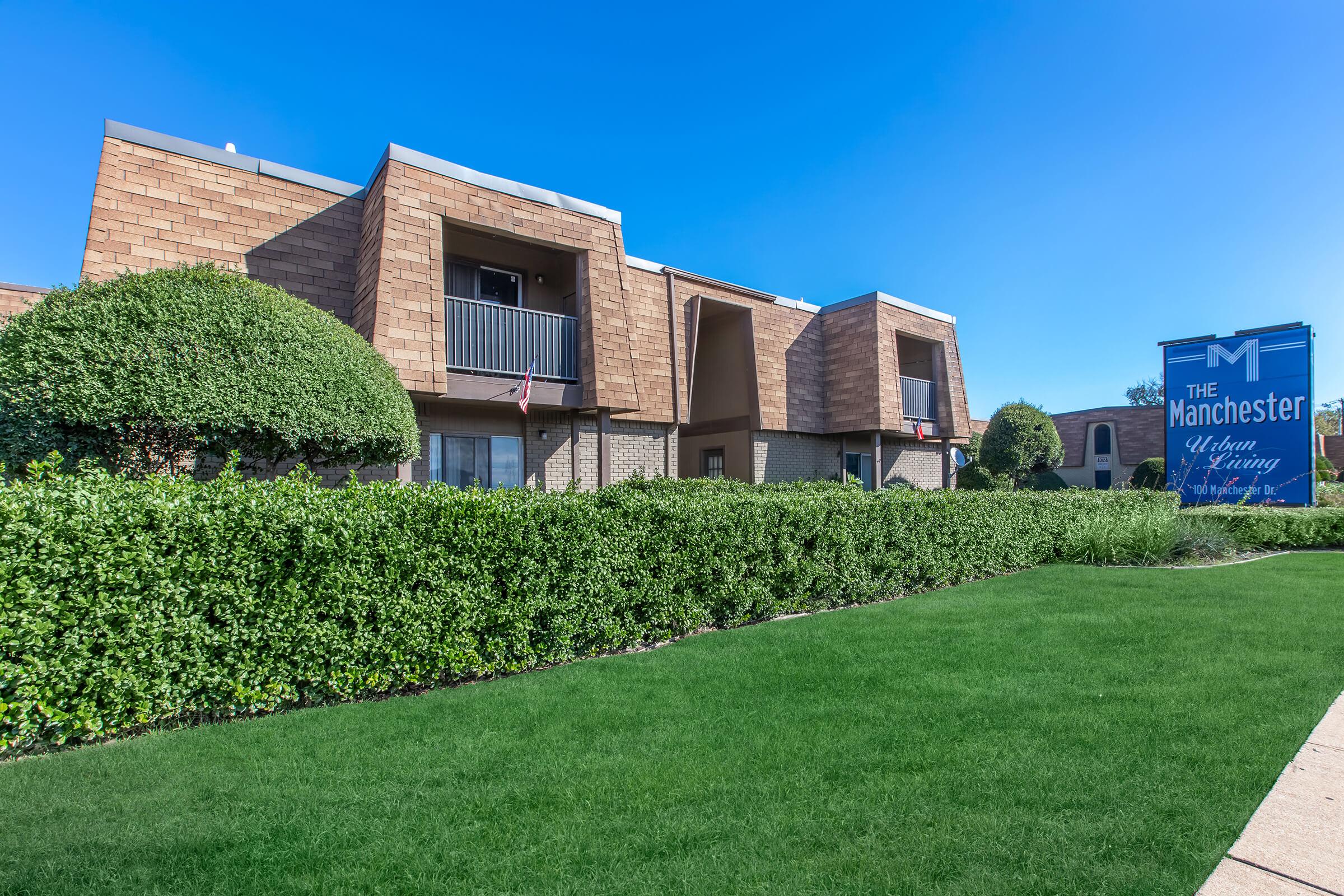 A sunny exterior view of an apartment community named "The Manchester." The building features a modern design with sloped roofs and balconies. Lush green landscaping with neatly trimmed hedges and a lawn adds to the appeal. A bright blue sign indicates the name of the complex.