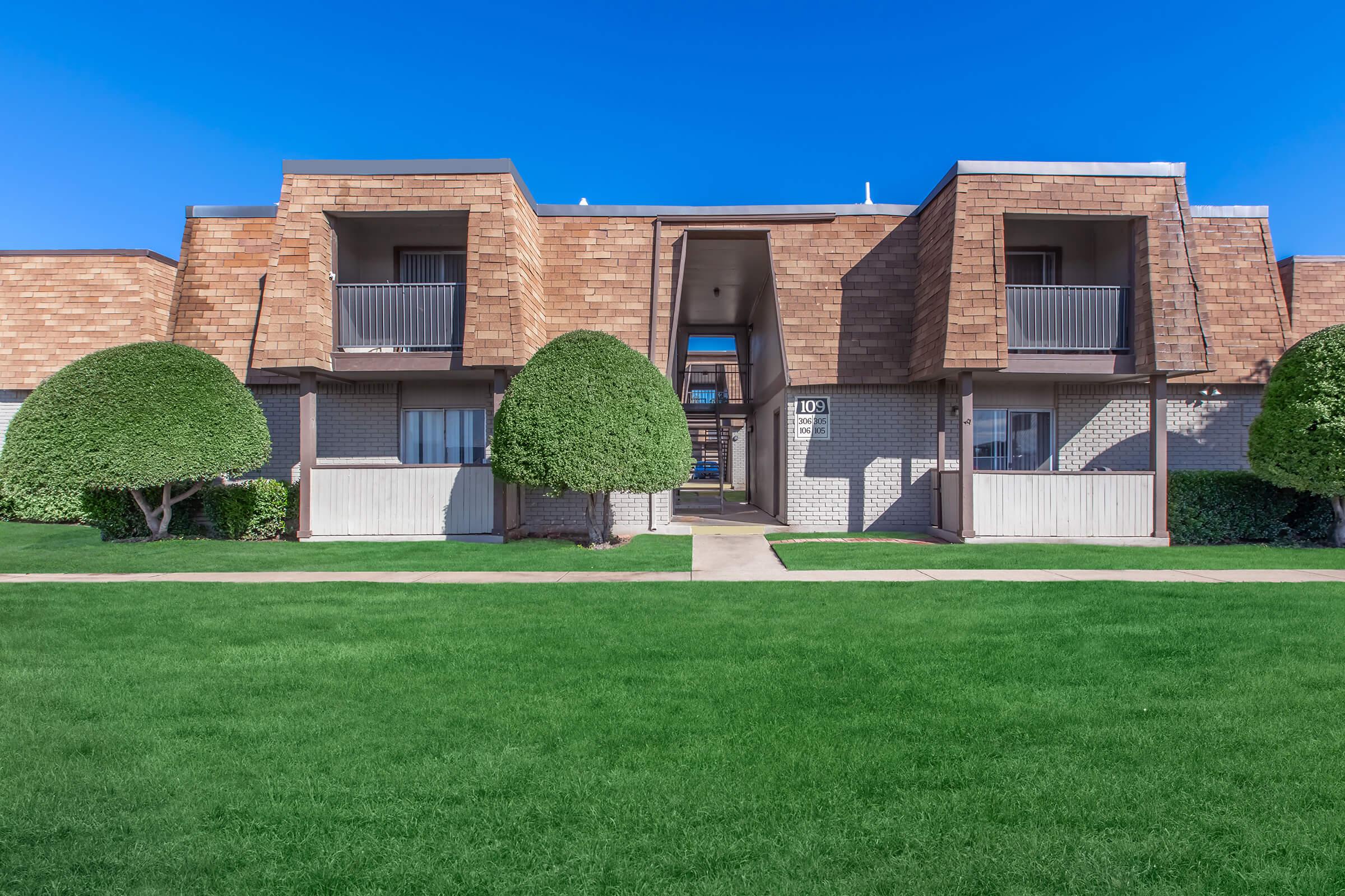 A two-story apartment building with a symmetrical facade. Each unit has a balcony with brown shingles on the roof. There are manicured round hedges and well-maintained green grass in the foreground, under a clear blue sky. The pathway in front leads to the entrance of the building.