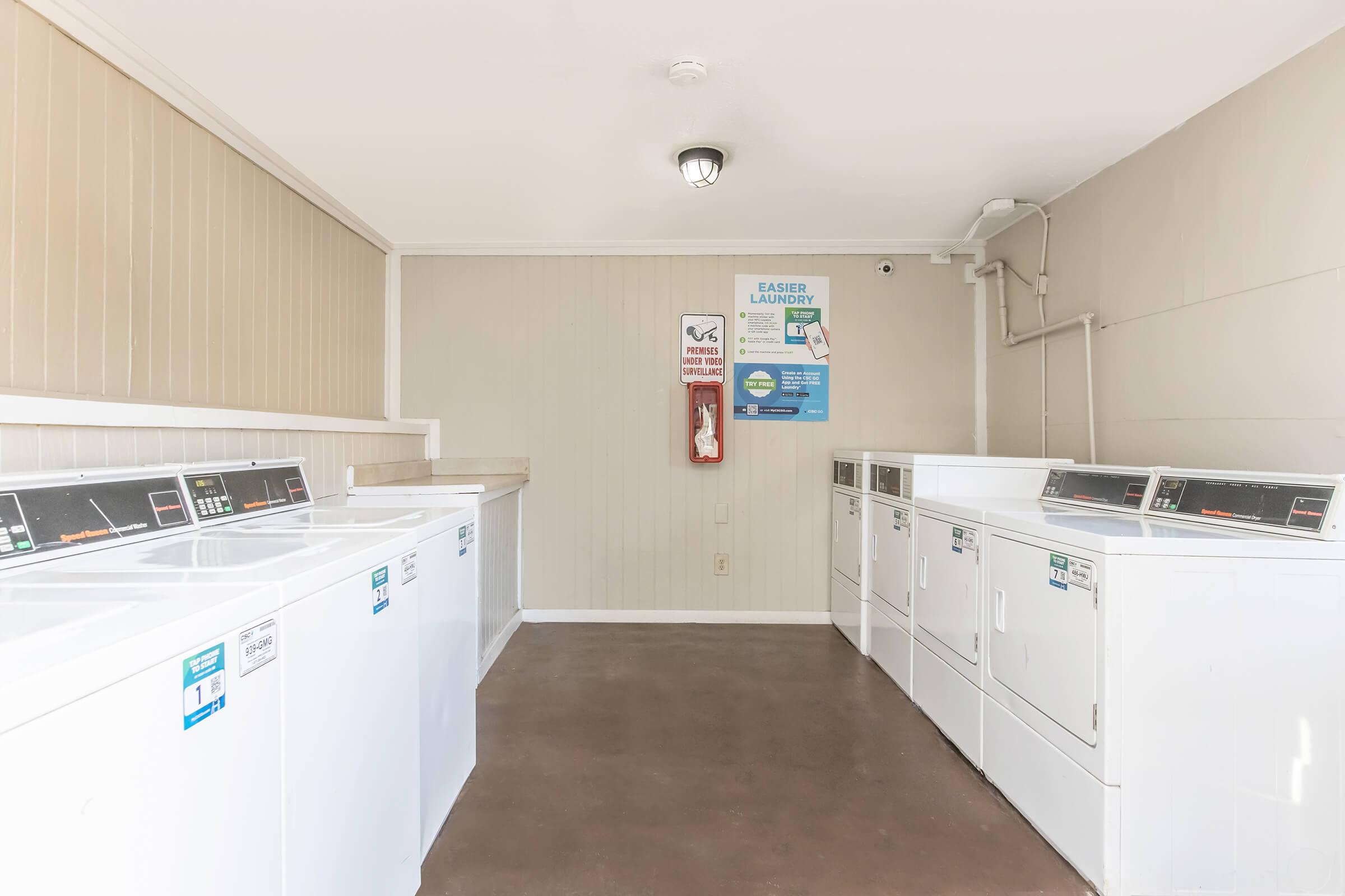 A clean, well-lit laundry room featuring several white washing machines and dryers arranged in a row. The walls are painted light beige, and there is a fire extinguisher mounted on the wall. A poster with laundry instructions is visible. The floor is a polished concrete.