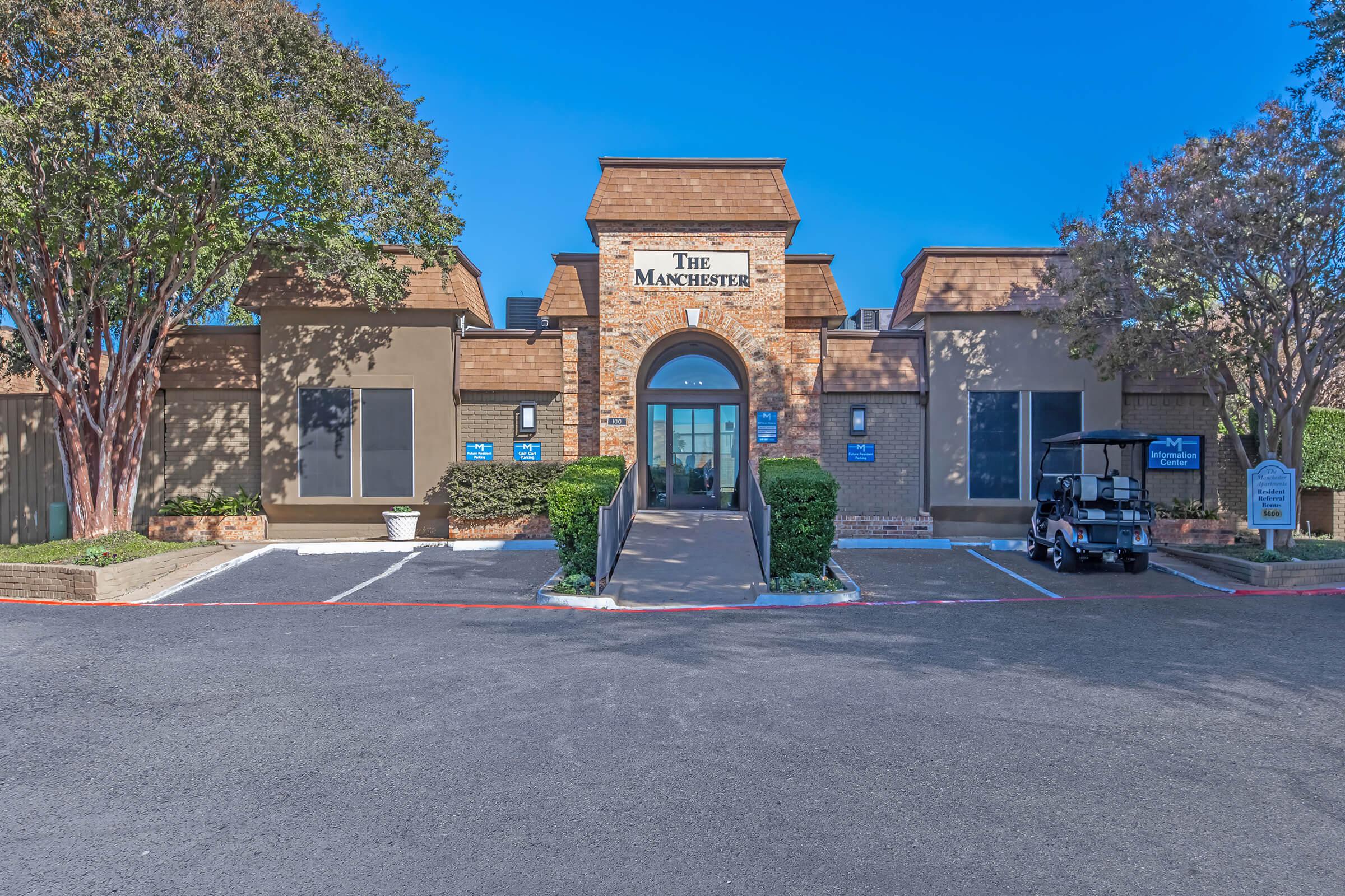 Exterior view of a building called "The Manchester," featuring a brick archway entrance and large windows. Surrounding landscaping includes trimmed bushes and trees. A golf cart is parked nearby, and the sky is clear and blue. The road in front is paved, with a few signage elements visible.