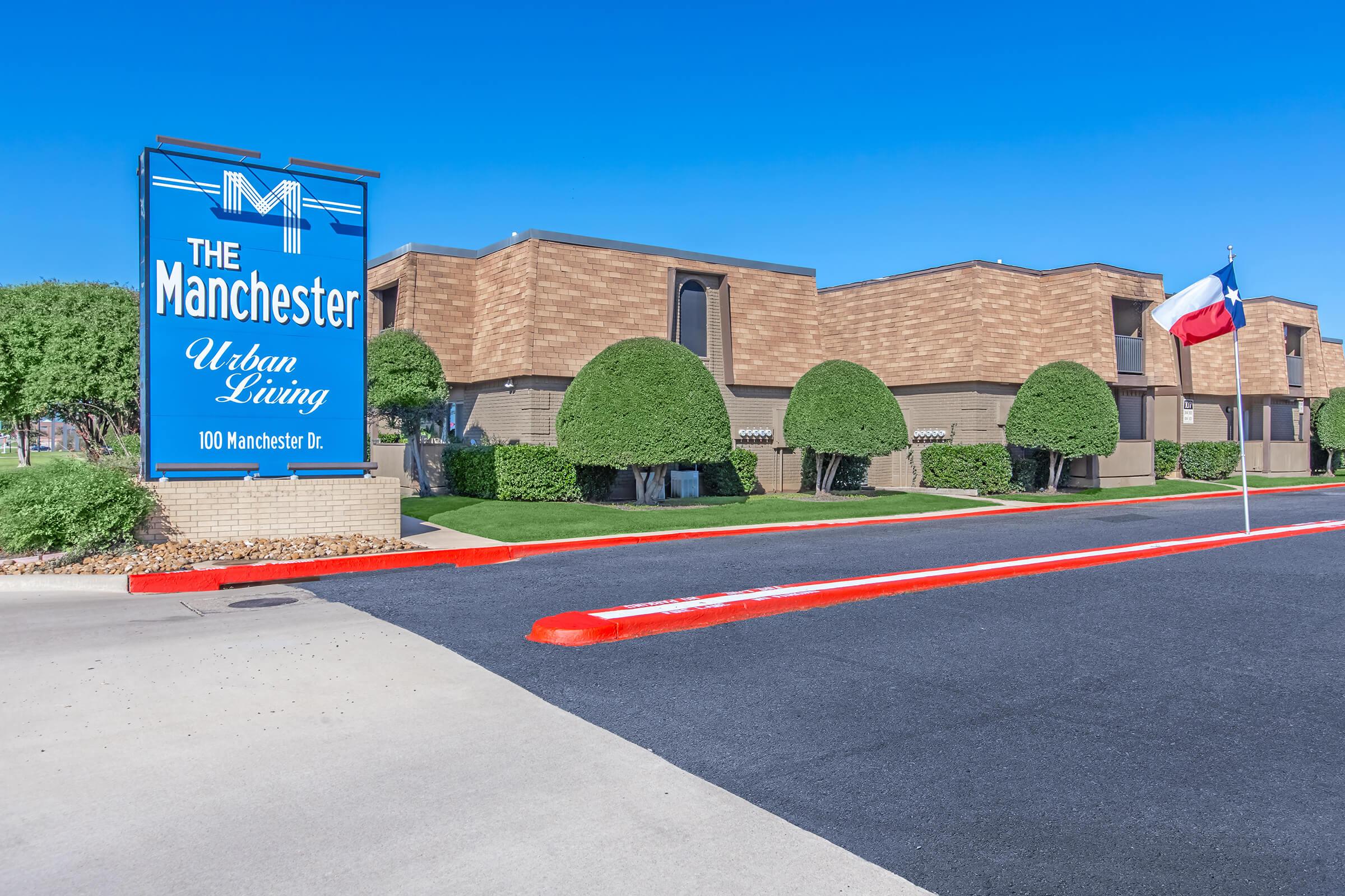 A sign for The Manchester Urban Living alongside a paved driveway. The building features a brick exterior and manicured landscaping with rounded hedges. A Texas flag is shown on a nearby flagpole. The clear blue sky creates a bright atmosphere. The address displayed is 100 Manchester Dr.