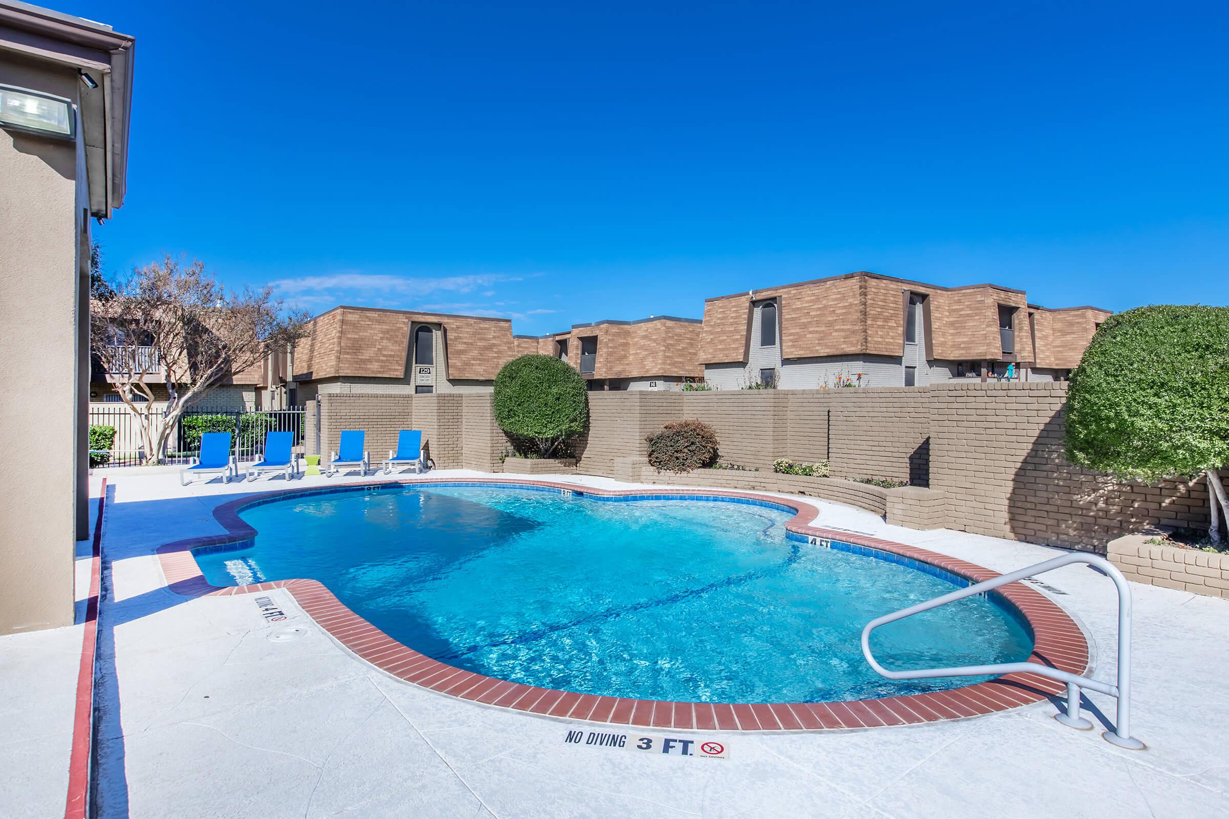 A clear swimming pool surrounded by a concrete deck and lounge chairs. In the background, there are apartments with brown roofs and neatly trimmed shrubs. The sky is bright blue, indicating a sunny day. A sign near the pool indicates a "No Diving" restriction, with a depth marker of 3 feet.