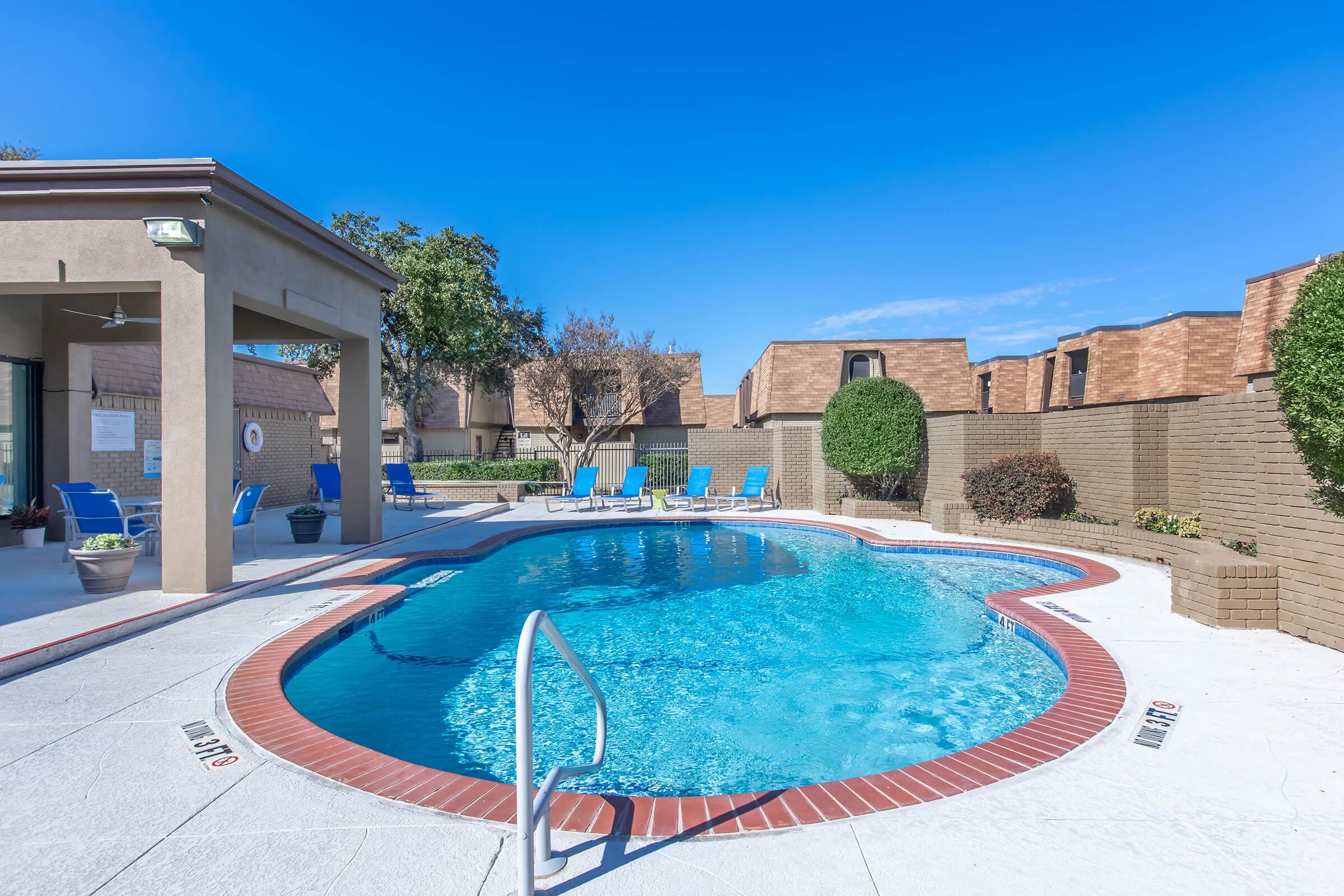 A clear swimming pool surrounded by lounge chairs under a bright blue sky. The pool is bordered by a beige tiled area and features landscaped greenery, including shrubs and trees. Nearby buildings have sloped roofs, typical of residential complexes.