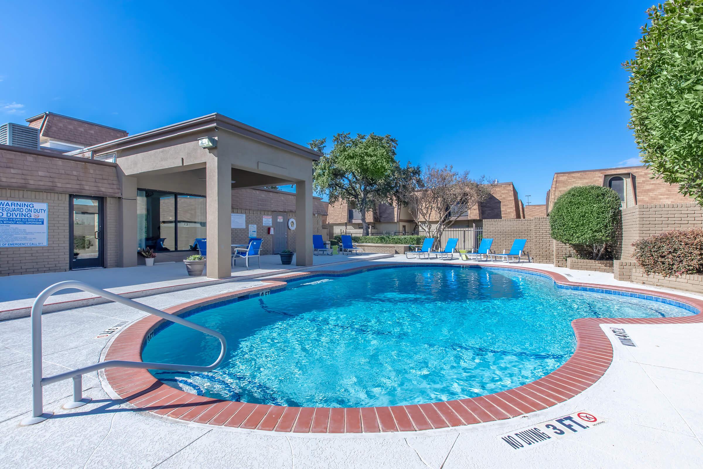 A clear blue swimming pool surrounded by lounge chairs, with a nearby building featuring large windows. The pool area is sunny, with a few trees and shrubs in the background. A sign is visible near the pool, and there's a gentle slope leading into the water. The overall scene conveys a relaxing outdoor atmosphere.