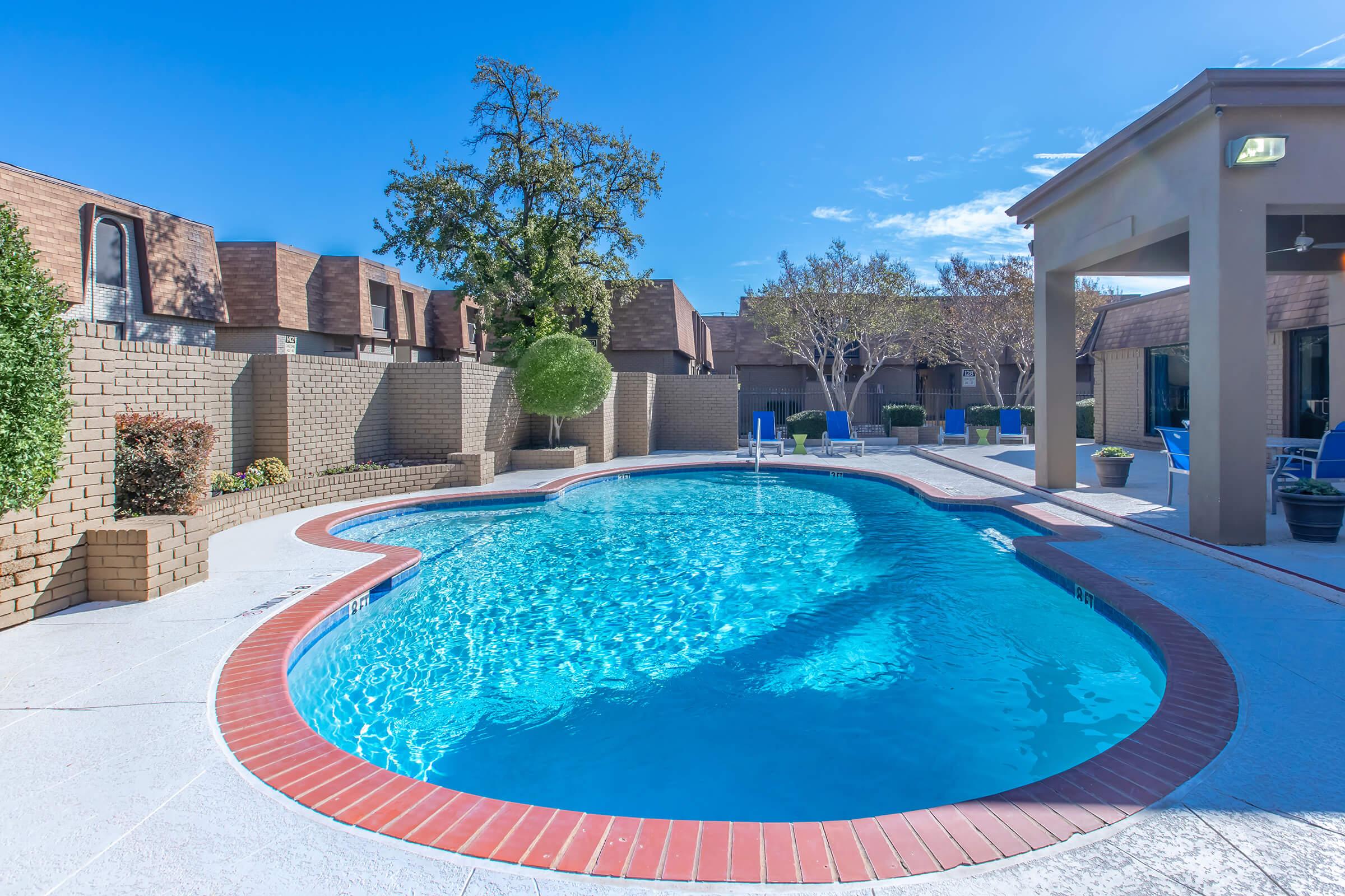 A clear swimming pool enclosed by a brick wall, surrounded by well-maintained landscaping. In the background, there are modern buildings with brown roofs and trees. A shaded seating area and lounge chairs are visible next to the pool under a bright blue sky.