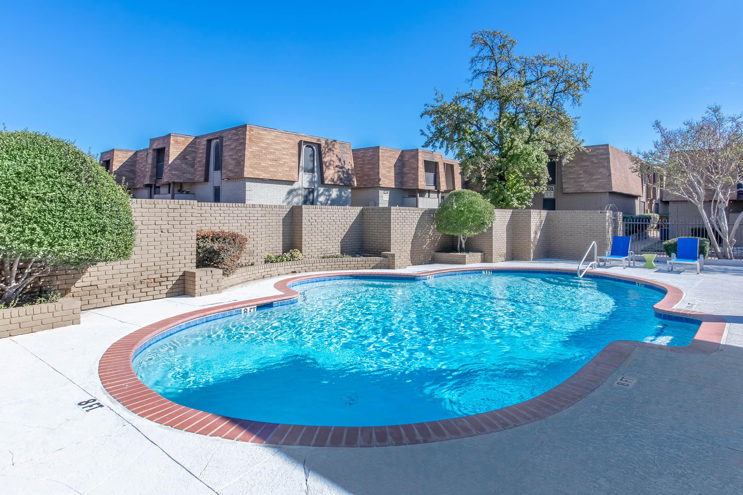 A clear, blue swimming pool surrounded by a beige brick wall and neatly trimmed bushes. In the background, there are several residential buildings with brown roofs. The sky is bright and blue, creating a sunny atmosphere. Poolside chairs are arranged near the edge of the pool.