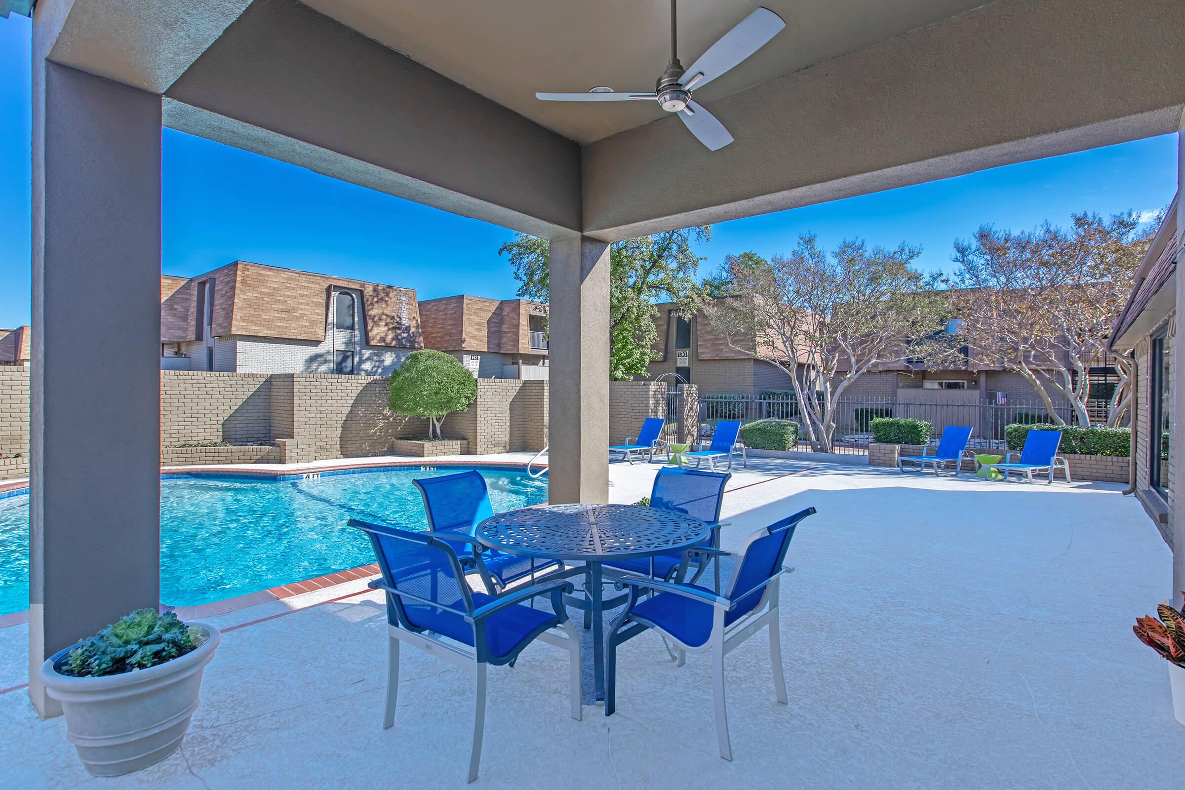 A sunny outdoor patio area featuring a round table with four blue chairs, a ceiling fan above, and a swimming pool in the background. Surrounding the pool are lounge chairs and landscaped greenery, set against a clear blue sky.